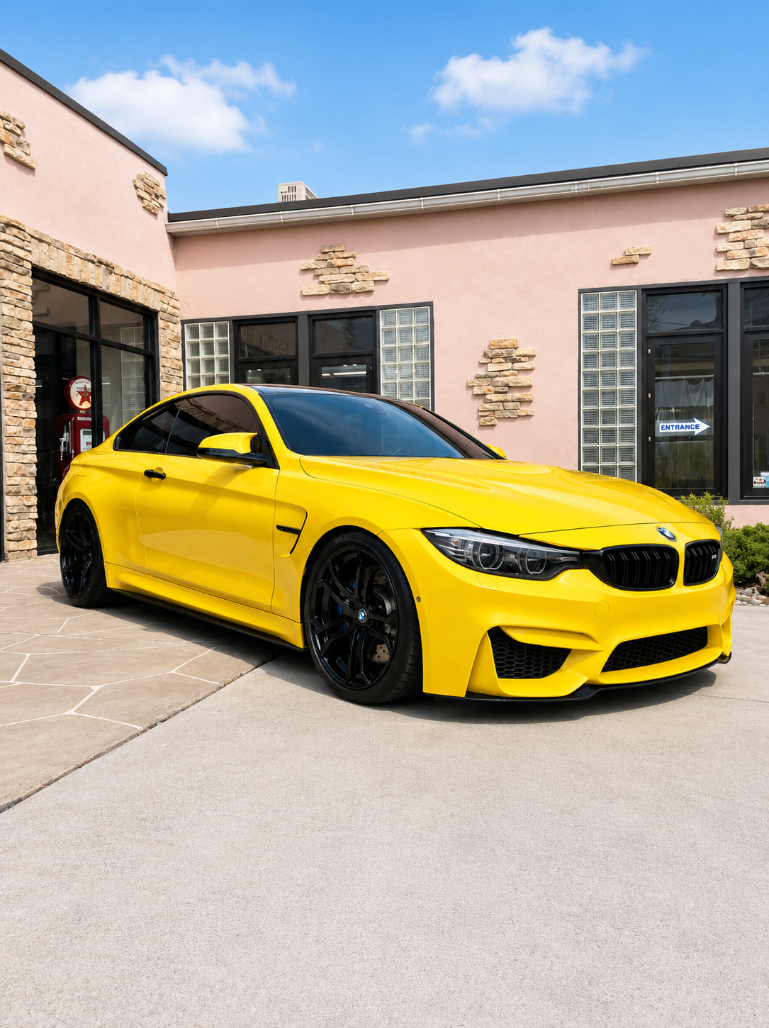 Yellow BMW sports car parked outside a modern building with pink walls and stone accents, under a partly cloudy blue sky.
