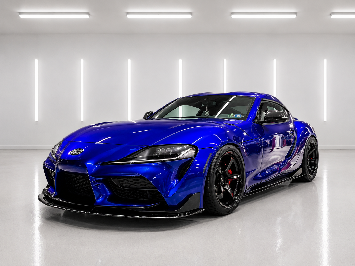 A blue sports car parked in a brightly lit, minimalist indoor space with a white floor, white walls, and vertical strip lights.