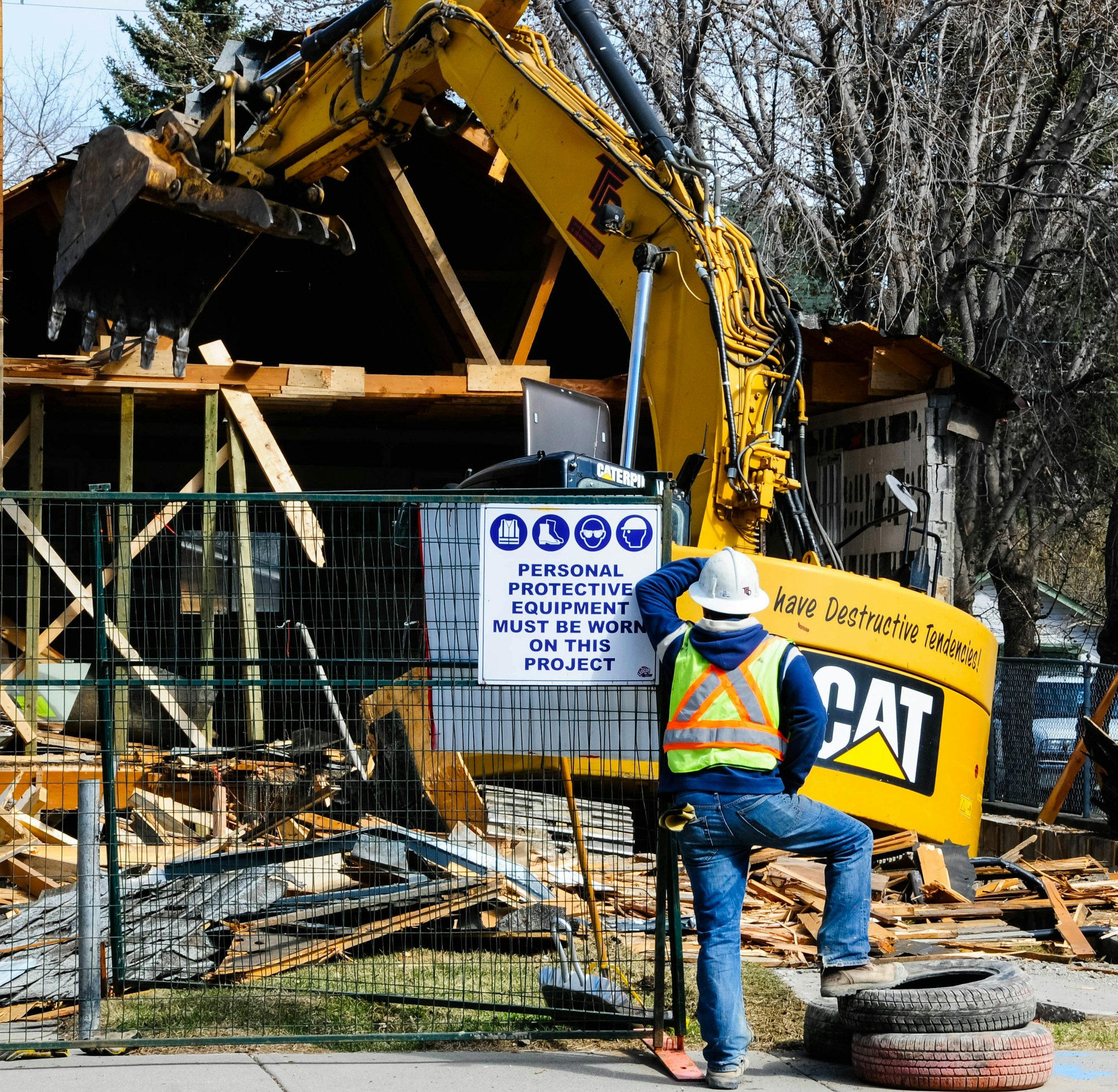 Construction worker wearing a yellow safety vest and white helmet working at a building site surrounded by broken wood, bricks, and debris, with a fence and a sign that indicates personal protective equipment must be worn.