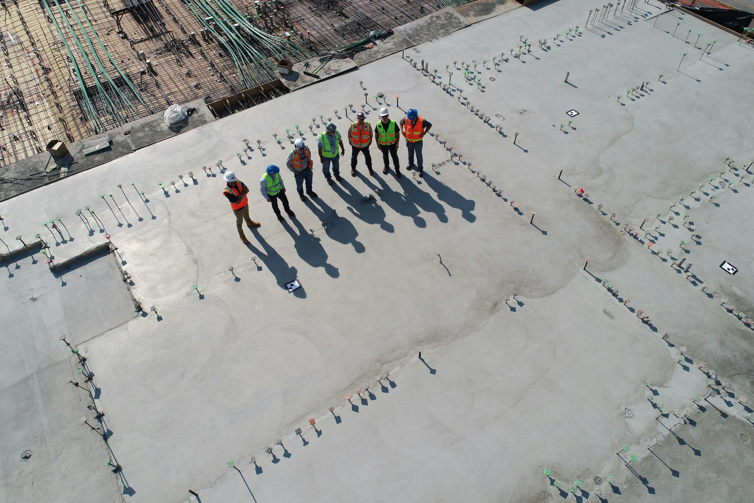 Six construction workers wearing safety vests and helmets standing on a concrete surface at a construction site, with rebar and construction materials around them.