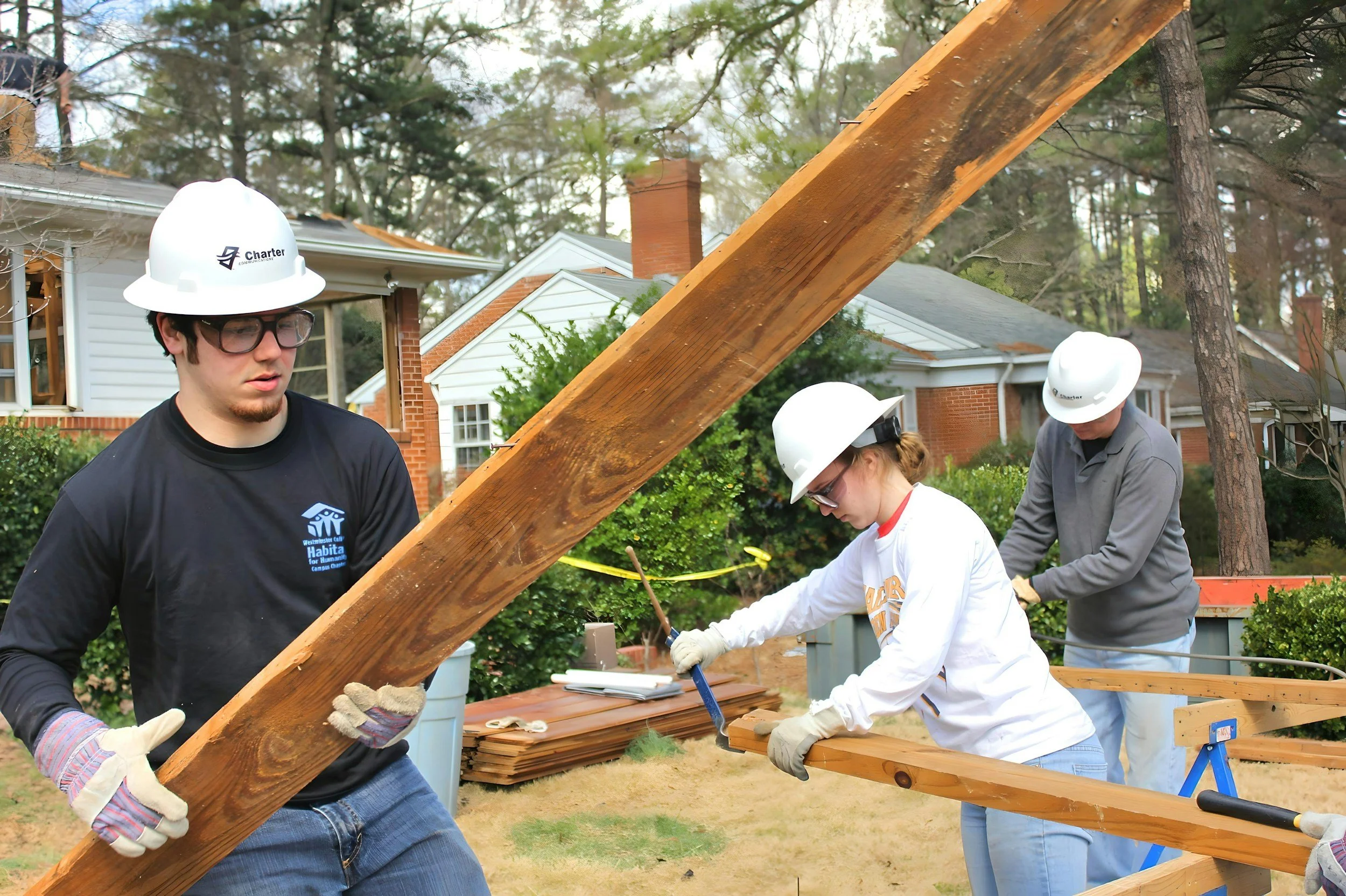 People working on a wooden structure outdoors, wearing white safety helmets and gloves, in a residential area with trees and houses in the background.