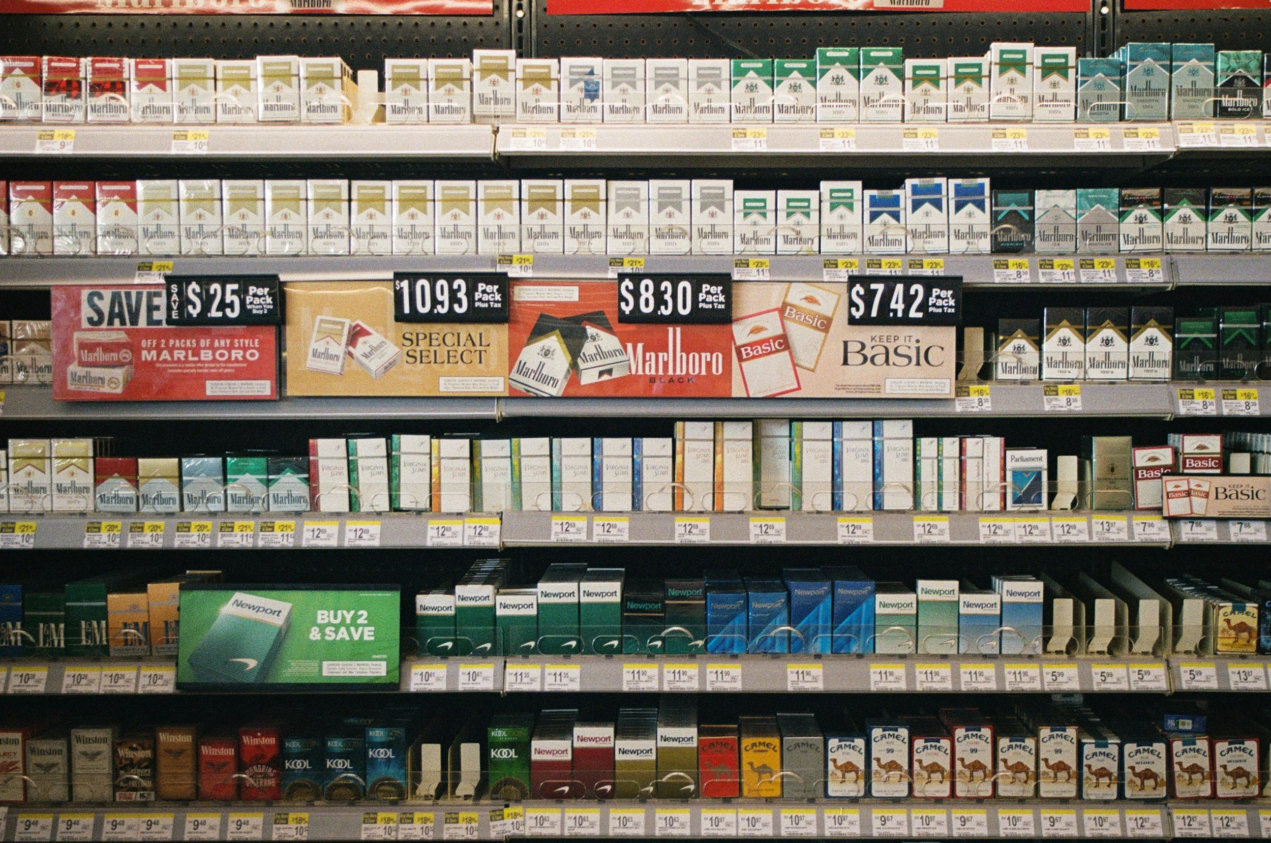 Shelf in a store displaying various brands and types of cigarettes, with prices and promotional signs for Marlboro cigarettes.