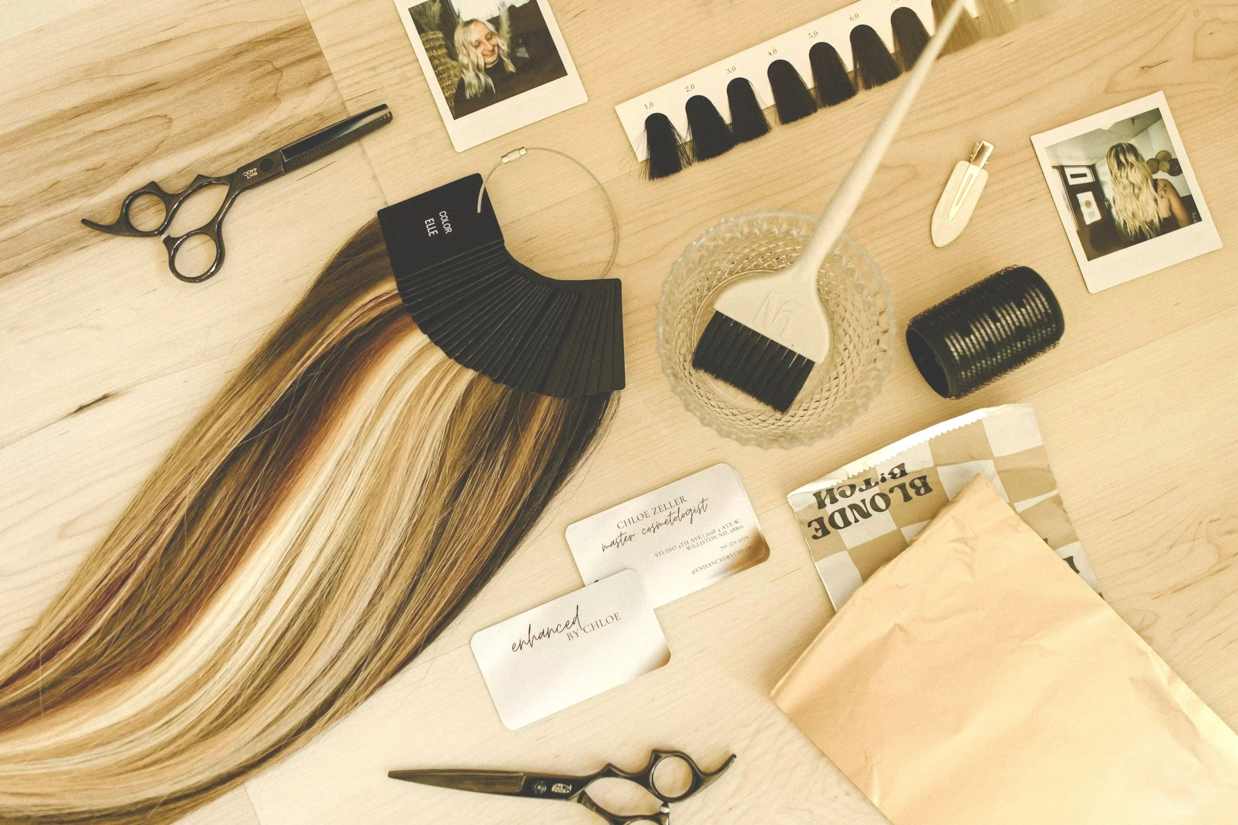 Hair color swatch samples, hair cutting scissors, hairbrush, Polaroid photos of a woman with blonde hair, business cards, hair color chart, hair extension sample, hair dye packet, hair roller, and a hair dye brush on a wooden table.