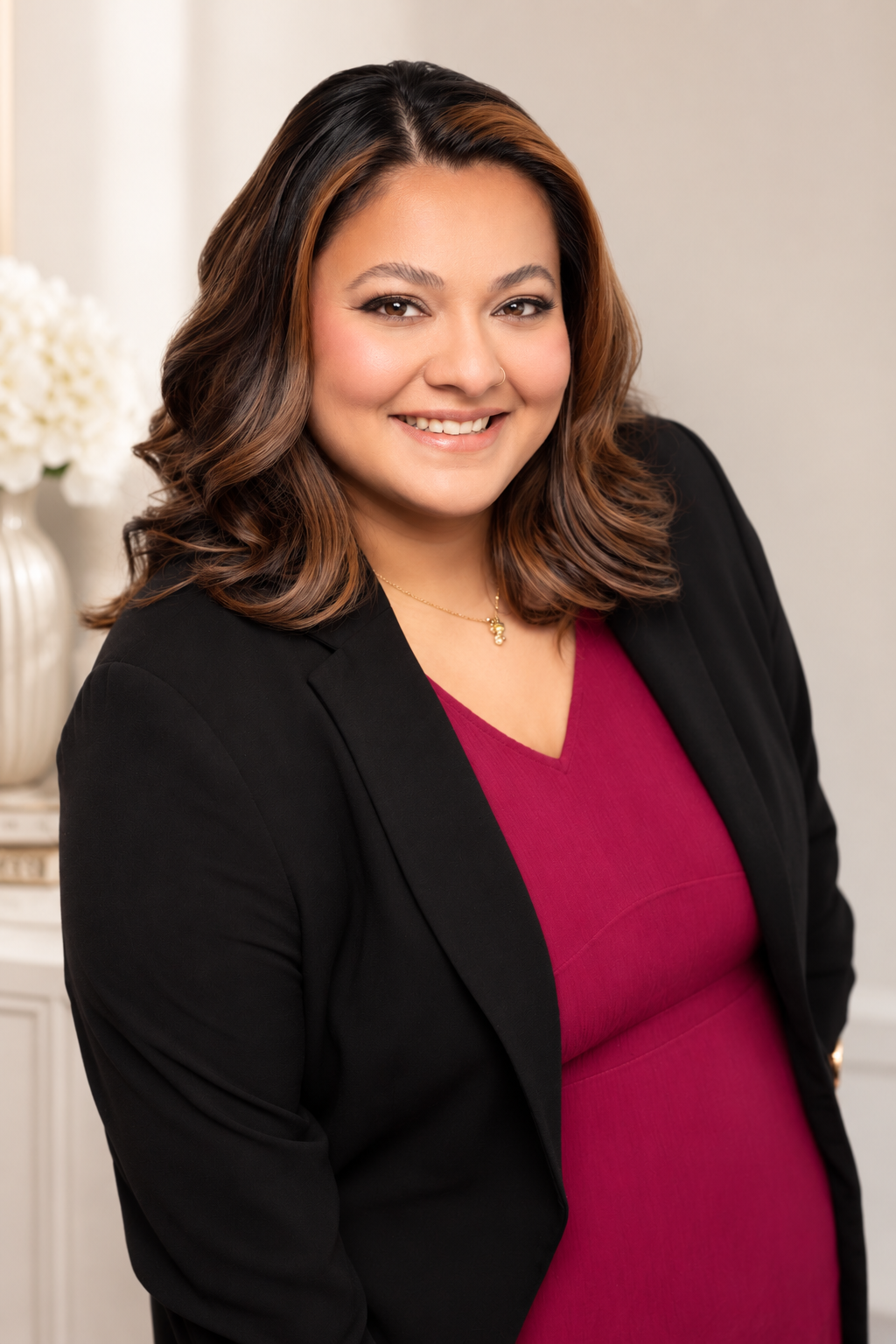 A woman with medium-length wavy brown hair with highlights, smiling, wearing a black blazer over a maroon top, and a gold necklace, standing in front of a neutral background with a vase of white flowers.