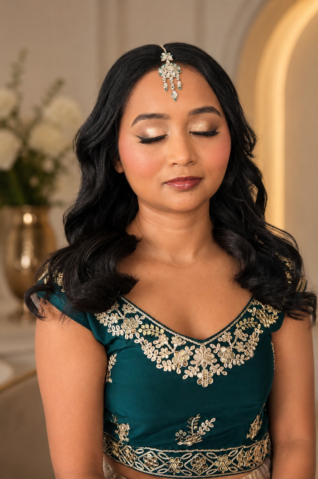 A woman with long black hair, closed eyes, and wearing makeup, adorns a teal dress with intricate gold embroidery. She has a decorative headpiece hanging over her forehead.