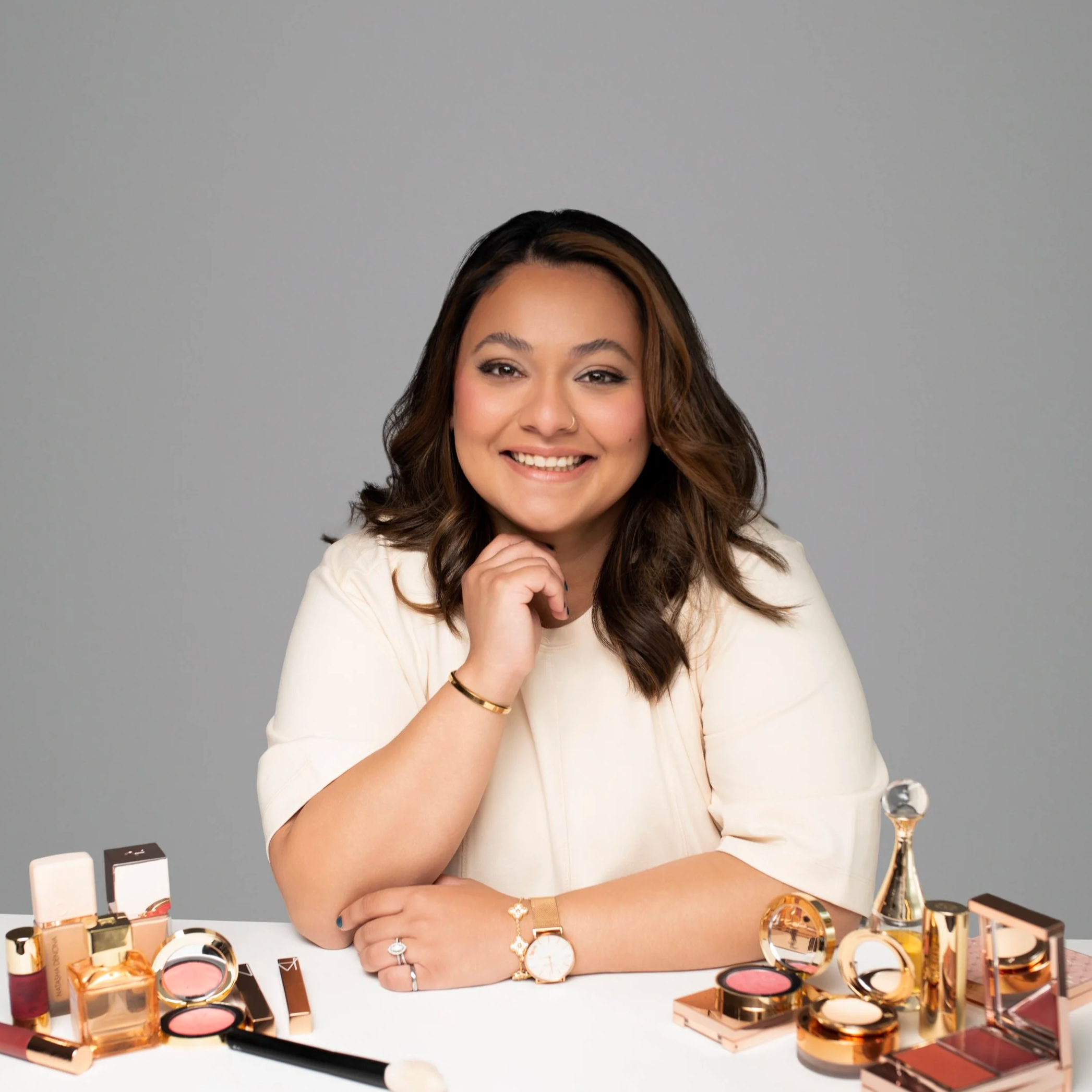 A woman with long, wavy brown hair and light brown skin smiling at a makeup display. She is wearing a white top and gold jewelry, including a watch, bracelet, rings, and a small nose ring. There are various makeup products such as blush, lip gloss, foundation, and brushes arranged on the white table in front of her, with additional makeup items visible in the background.