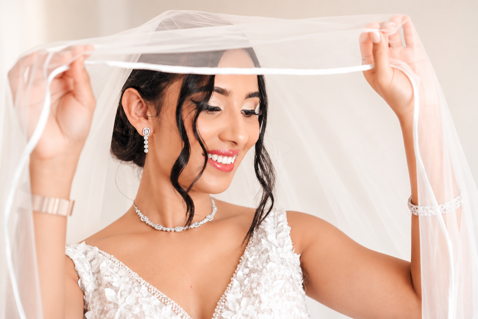 A bride smiling as she holds her wedding veil above her head, wearing a white wedding dress, pearl jewelry, and earrings.