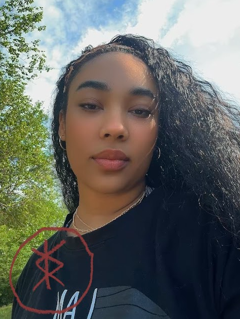 A selfie of a young woman outdoors with trees and blue sky, wearing a black shirt and gold chain necklace, with long curly hair and a pristine look to the camera.