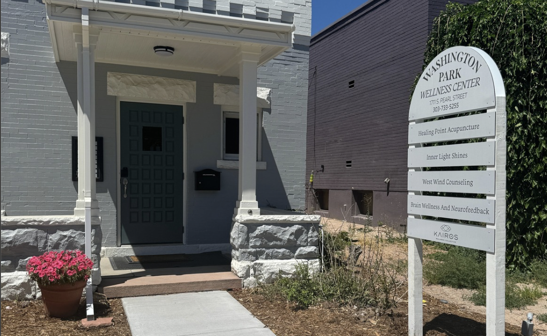 The entrance of Washington Park Wellness Center with a gray door, white brick facade, a flowerpot with pink flowers, and a white sign listing services like acupuncture and counseling.