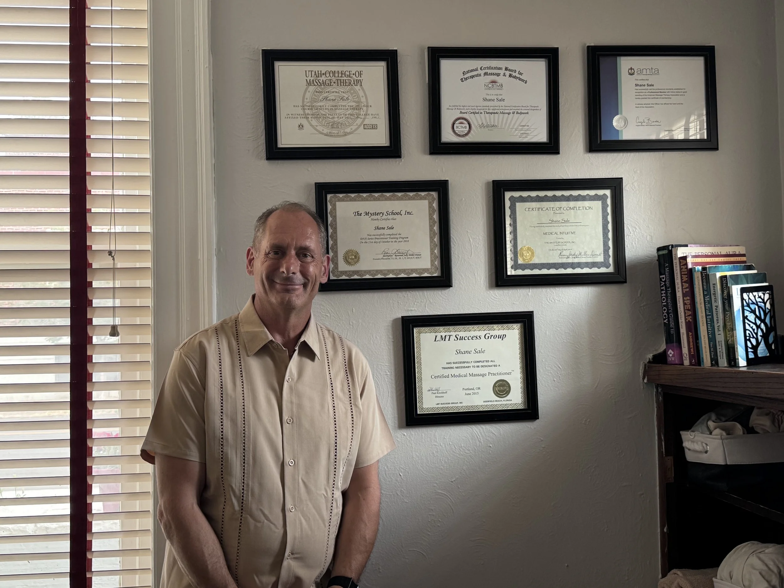Shane Sale, LMT smiling standing in front of a wall with multiple framed certificates and diplomas. There are books on a shelf to his right and a window with blinds to his left.