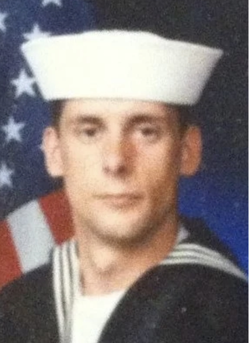 A young man in a U.S. Navy sailor uniform, wearing a white sailor hat, in front of an American flag background.