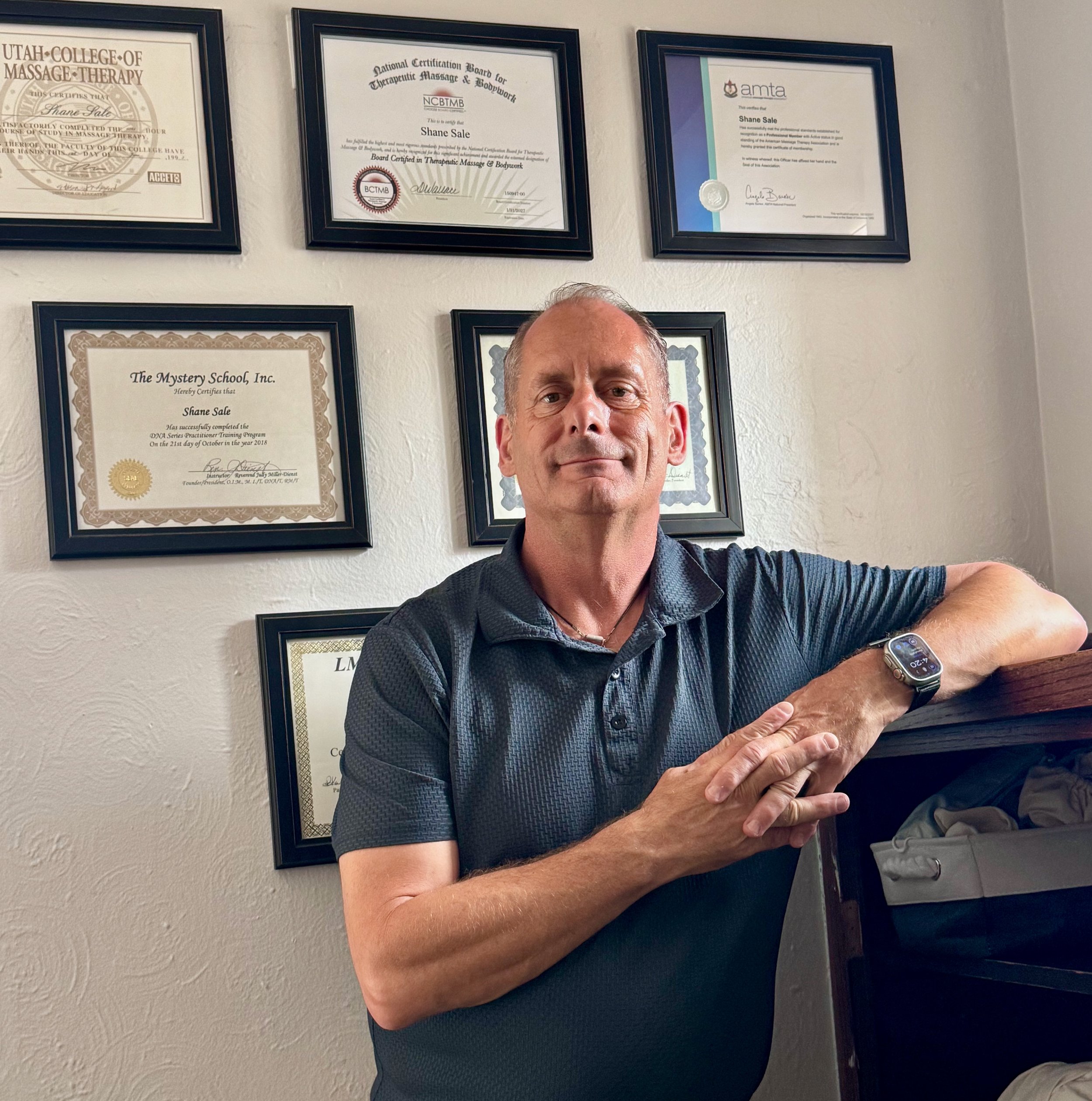 A middle-aged man with short hair, wearing a dark short-sleeved shirt and a wristwatch, taking a selfie in front of a wall decorated with framed certificates and diplomas.