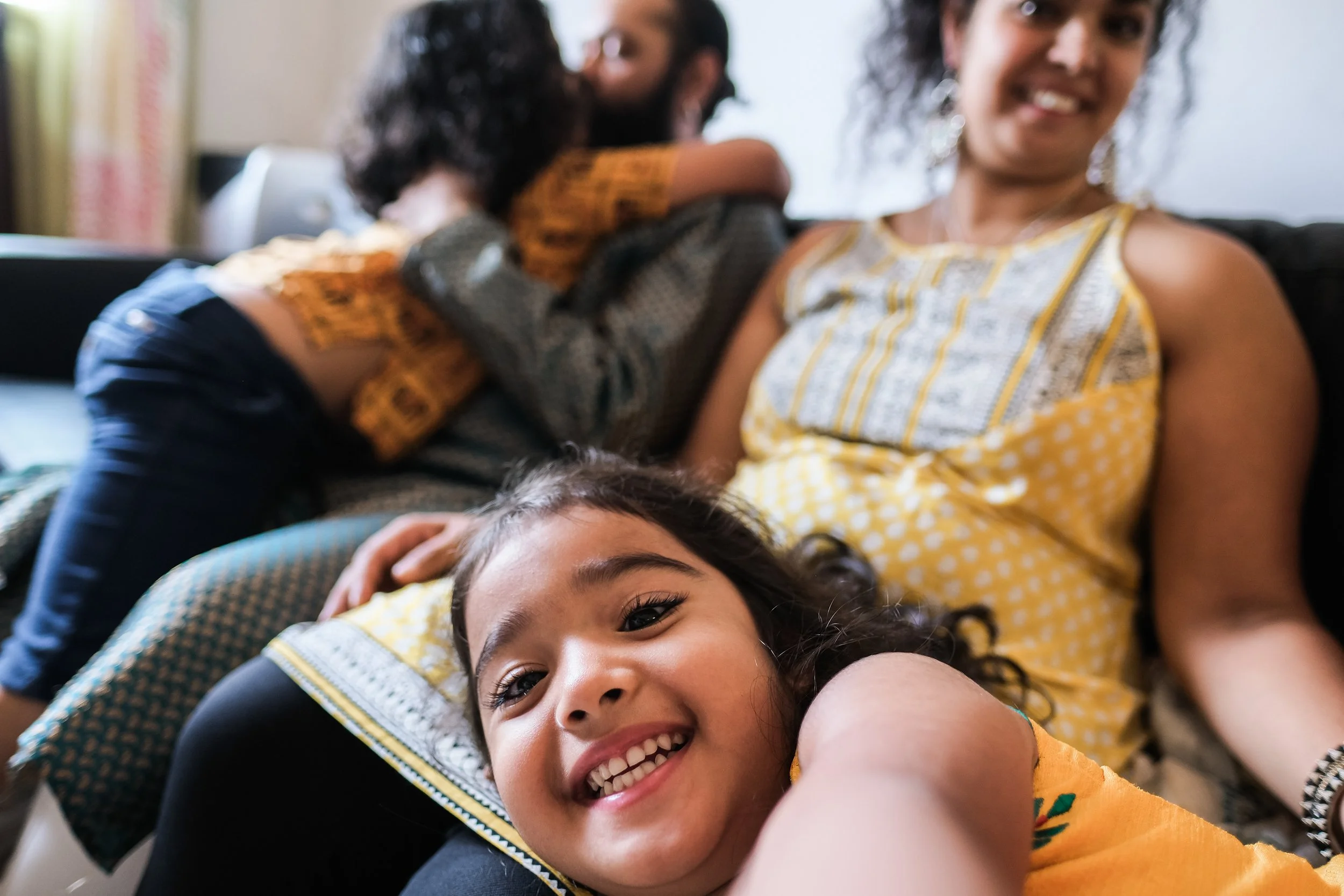 Three young women lying on the floor, smiling and taking selfies together. One woman in the foreground has curly hair and is wearing a yellow shirt with black patterns. The woman in the middle is hugging the woman on the left, and the woman on the right is smiling at the camera.