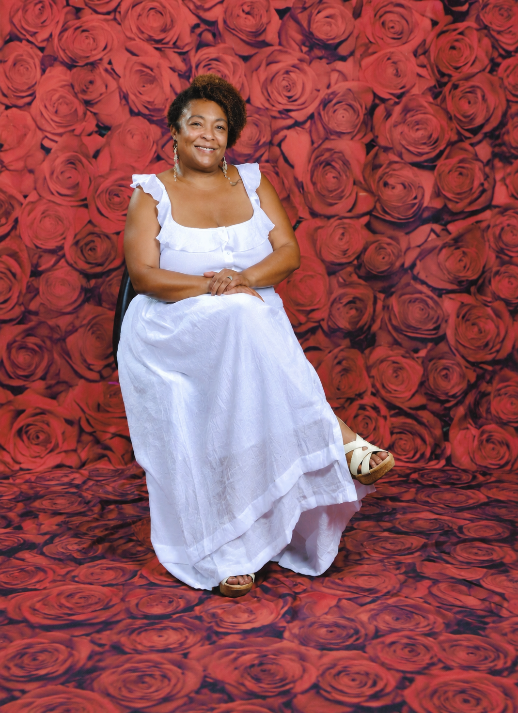 Angeleah sitting on a chair in front of a backdrop of red roses, wearing a white dress and white sandals, smiling at the camera.