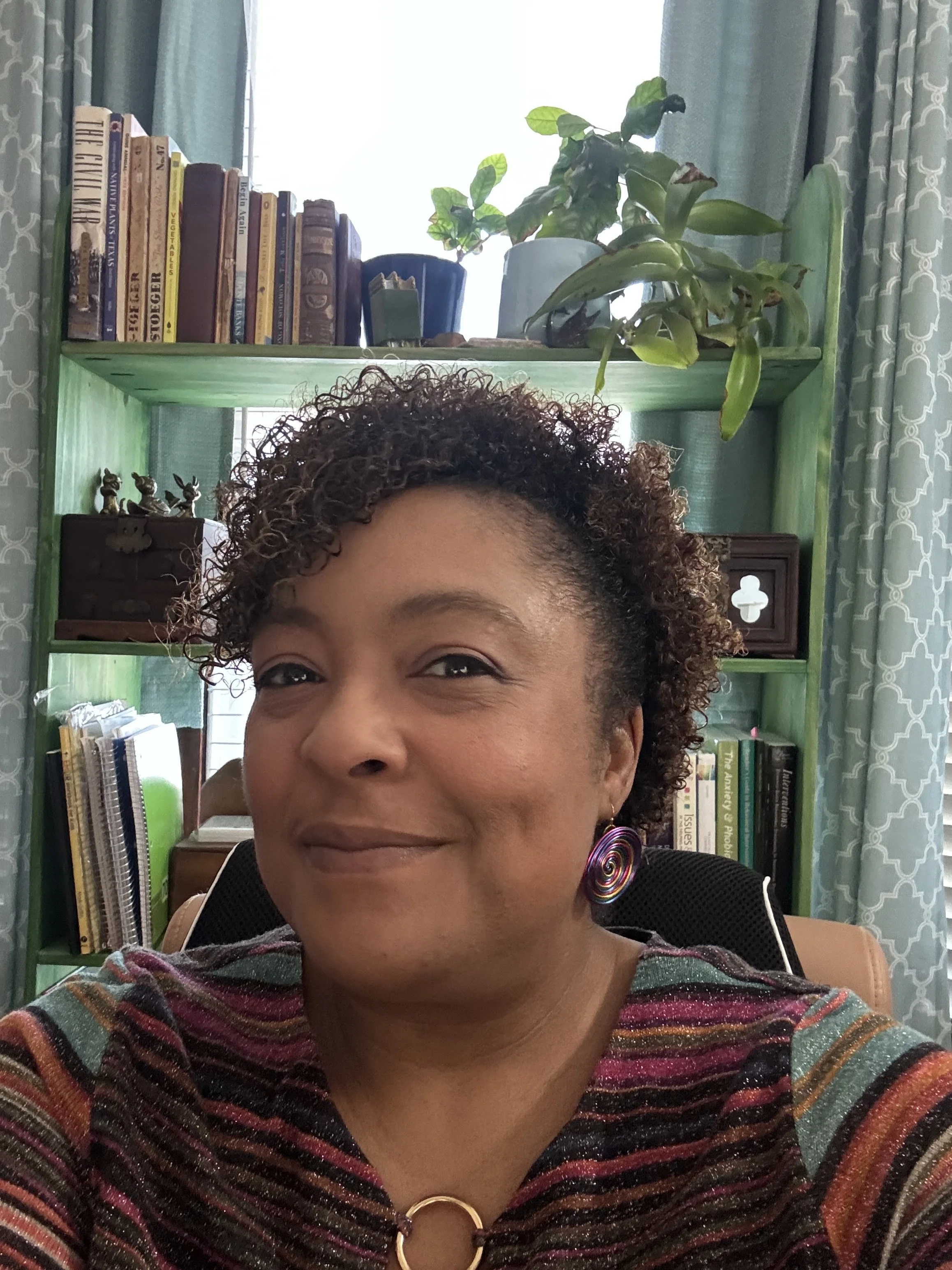 A woman with curly hair, wearing a colorful striped top and spiral earrings, smiling in a room with shelves filled with books and decorative items, and curtains with a patterned design.