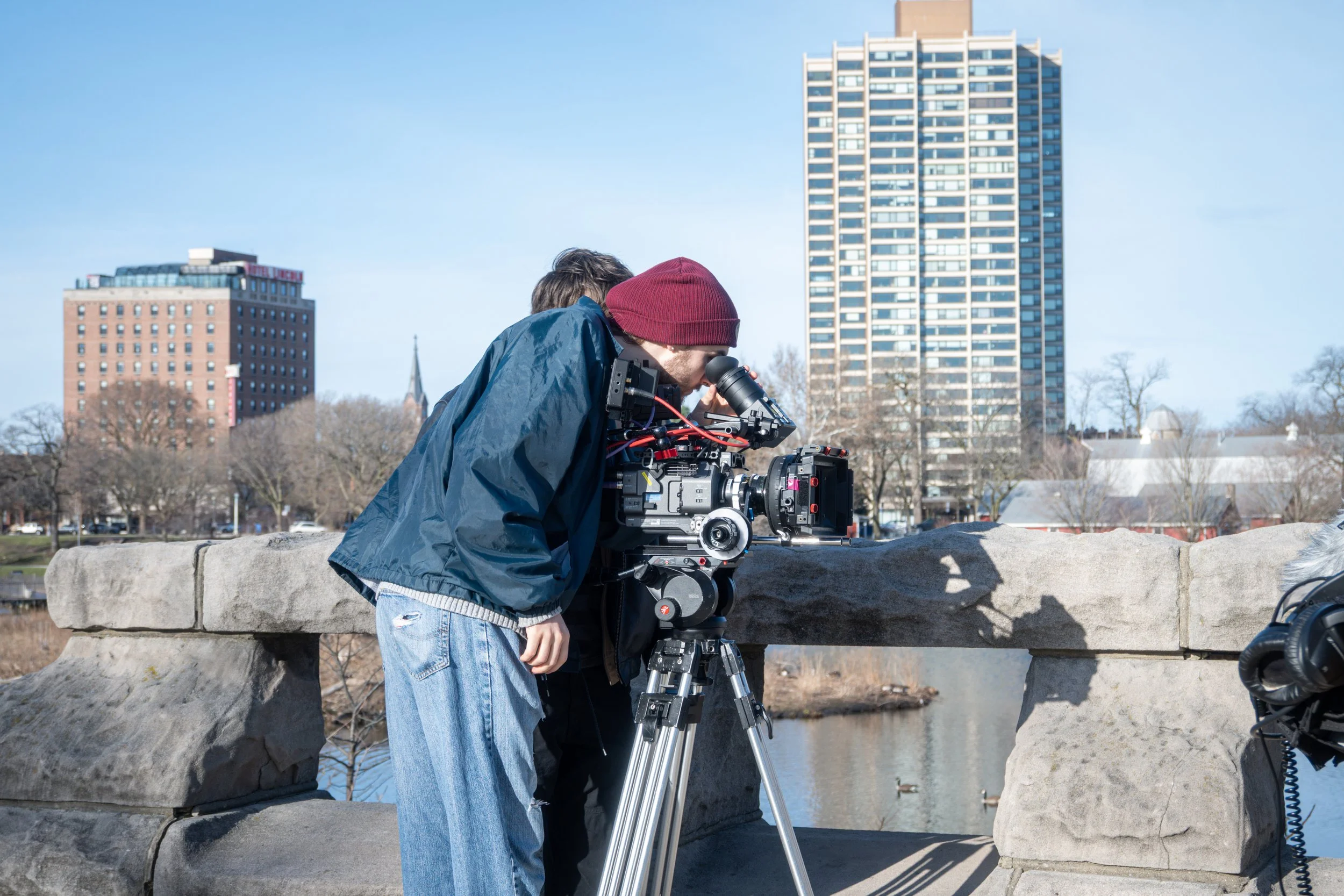 A person wearing a red beanie and a blue jacket looking into a camera on a tripod outdoors with city buildings and a park in the background.