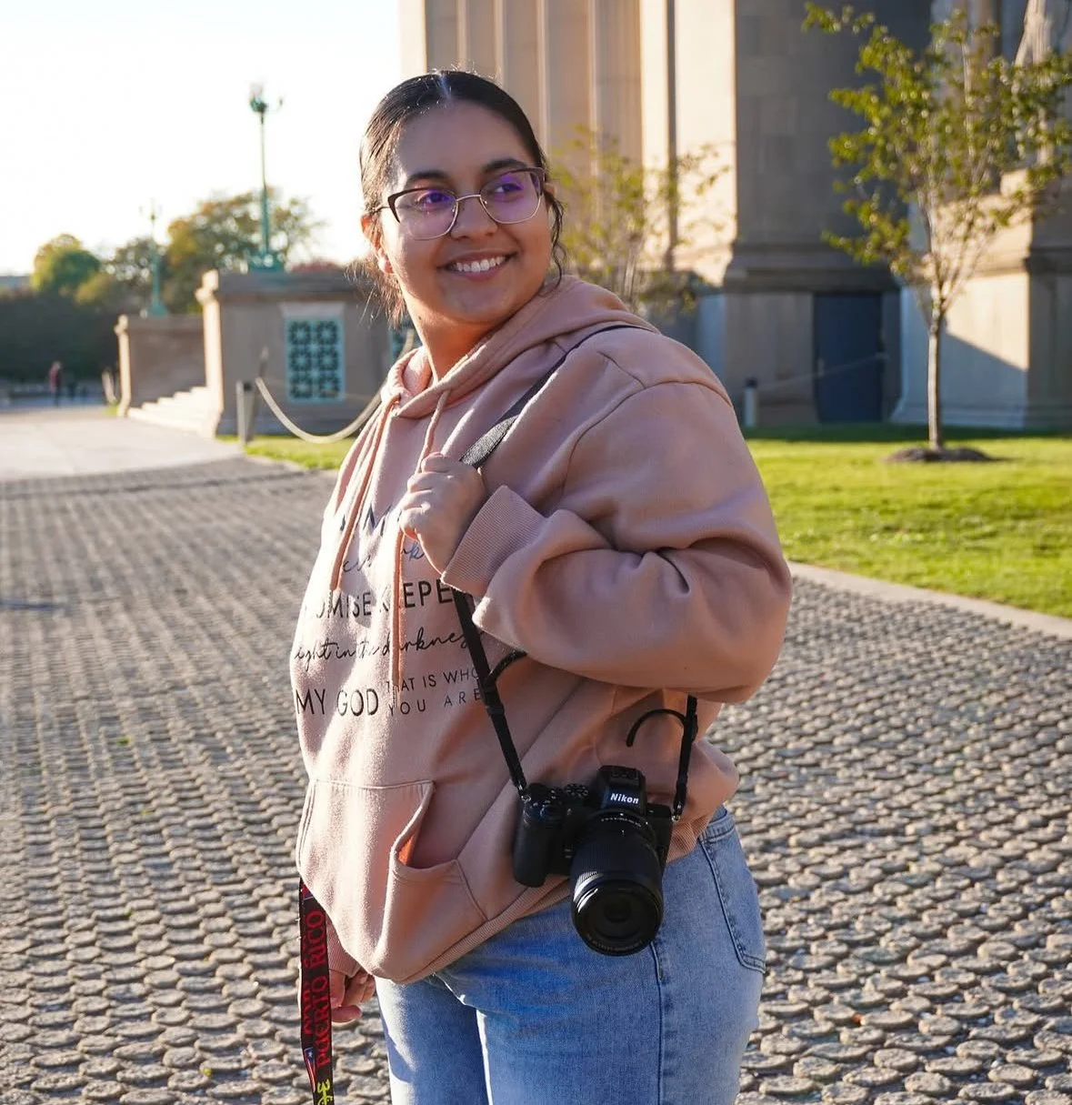 A smiling young woman with glasses and dark hair in a ponytail, wearing a pink hoodie and light blue jeans, holding a Nikon camera around her neck and standing outdoors near a building with trees and a cobblestone walkway.