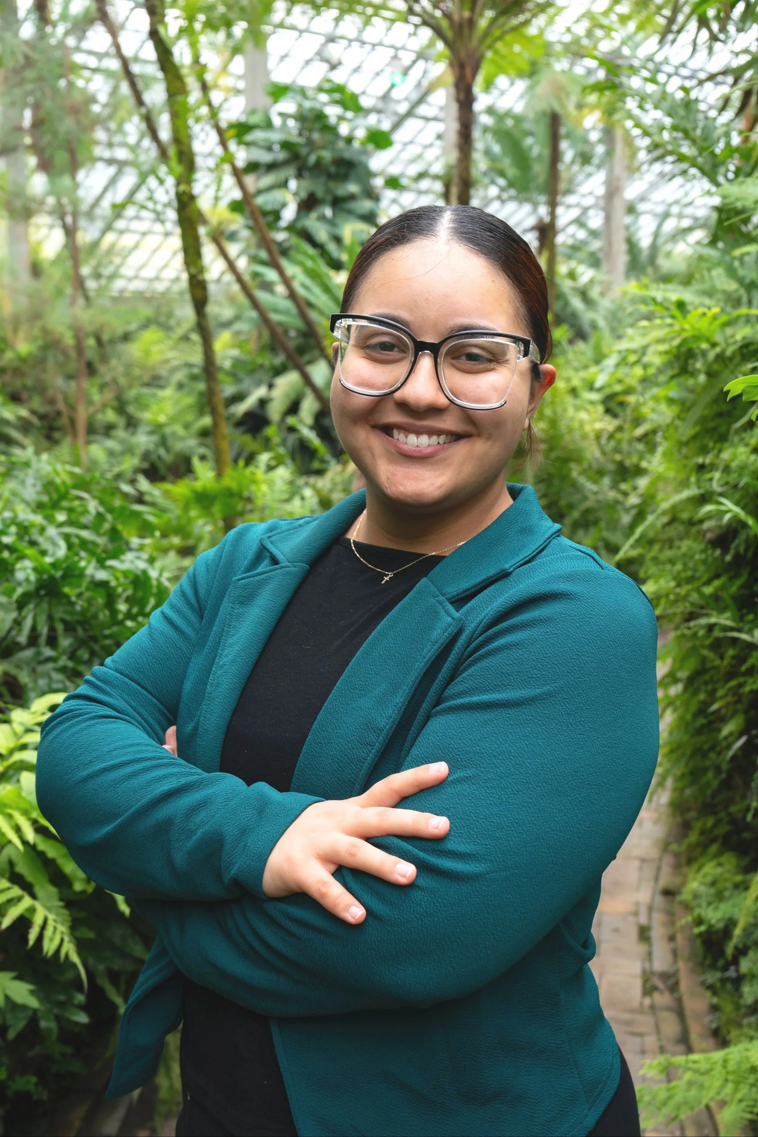 A woman with dark hair pulled back, wearing glasses, a black shirt, and a teal jacket, smiling with arms crossed, standing in a lush green indoor garden.