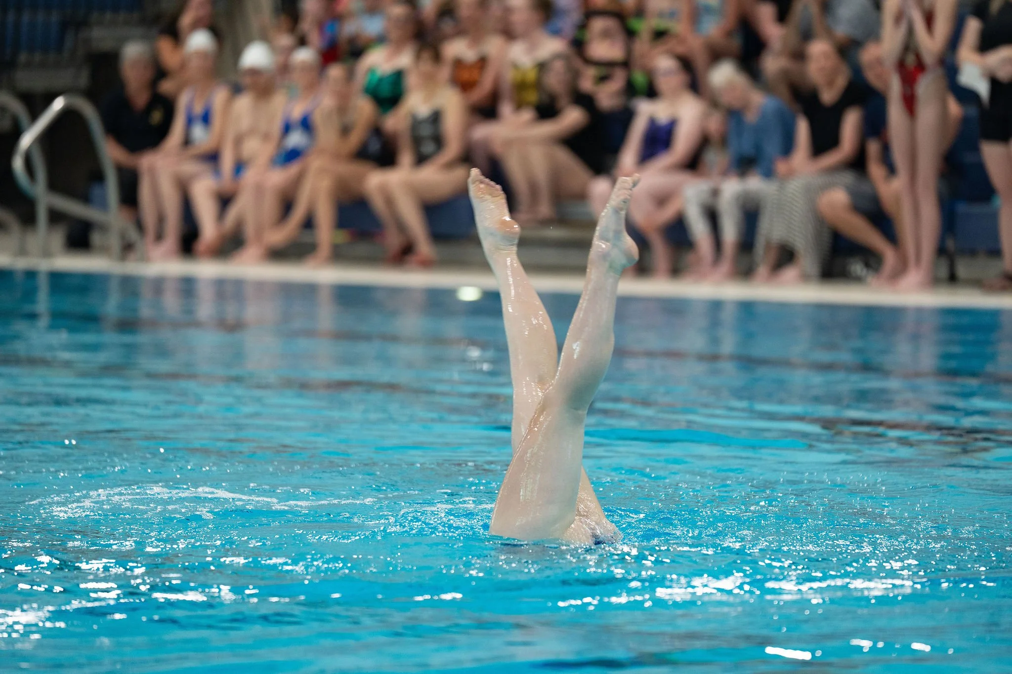 Swimmer is upside down in the pool, with legs and feet exposed out of the water, and other swimmers sitting on the pool deck watching in the background.
