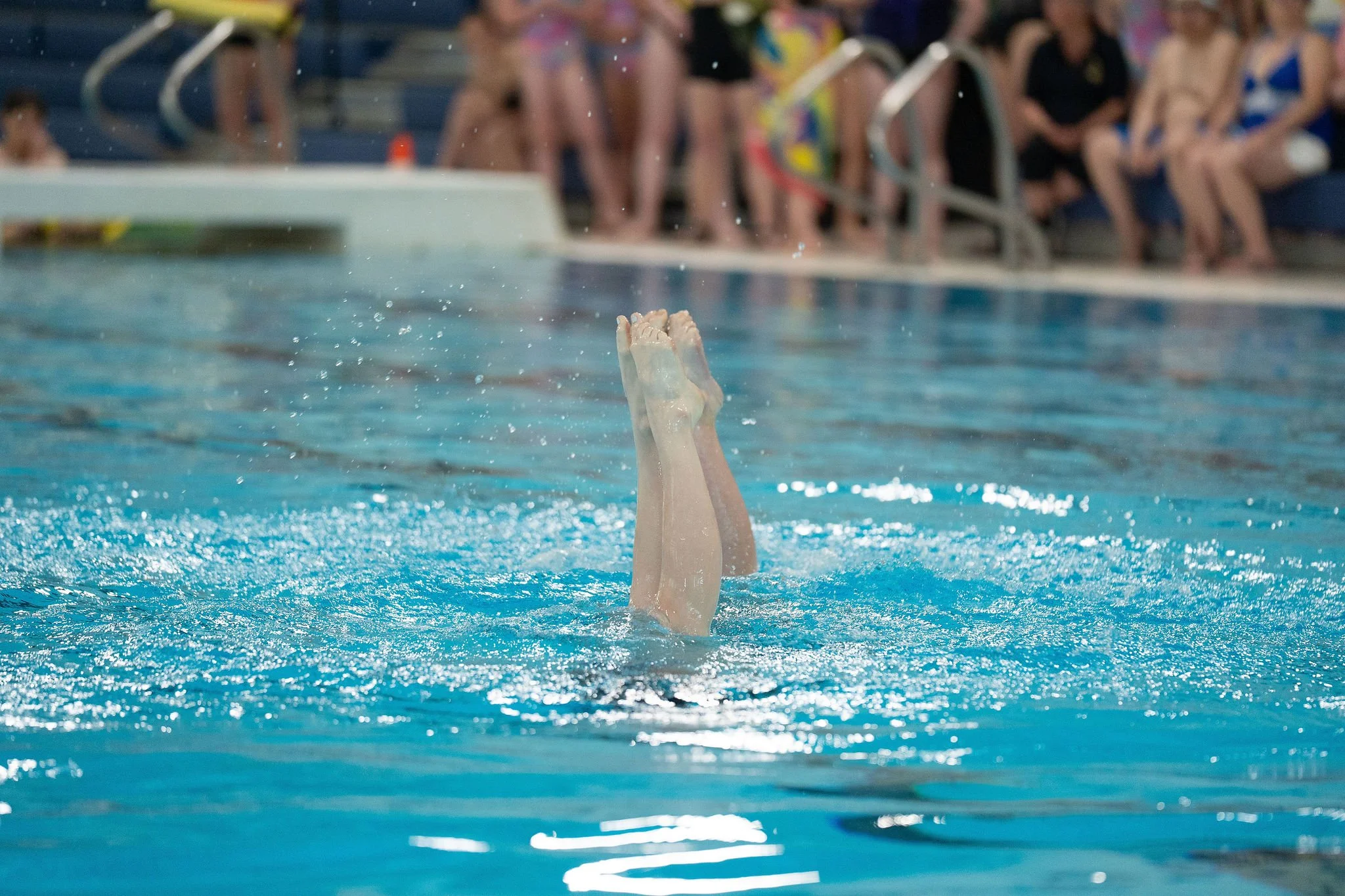Swimmer is upside down in the pool, with legs and feet exposed out of the water, and other swimmers sitting on the pool deck watching in the background.