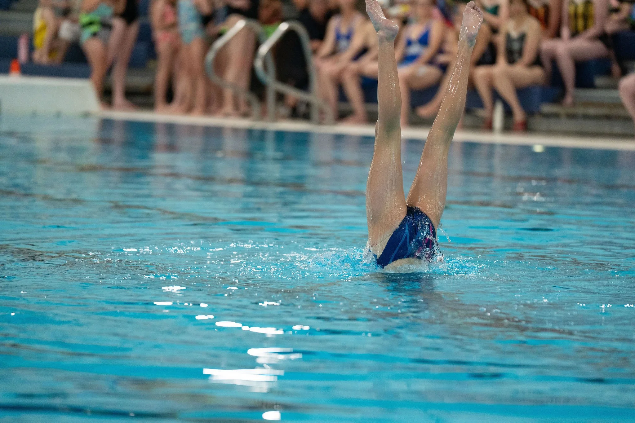 Swimmer performing a Duet acrobatic in a swimming pool during a competition, with an audience watching in the background.
