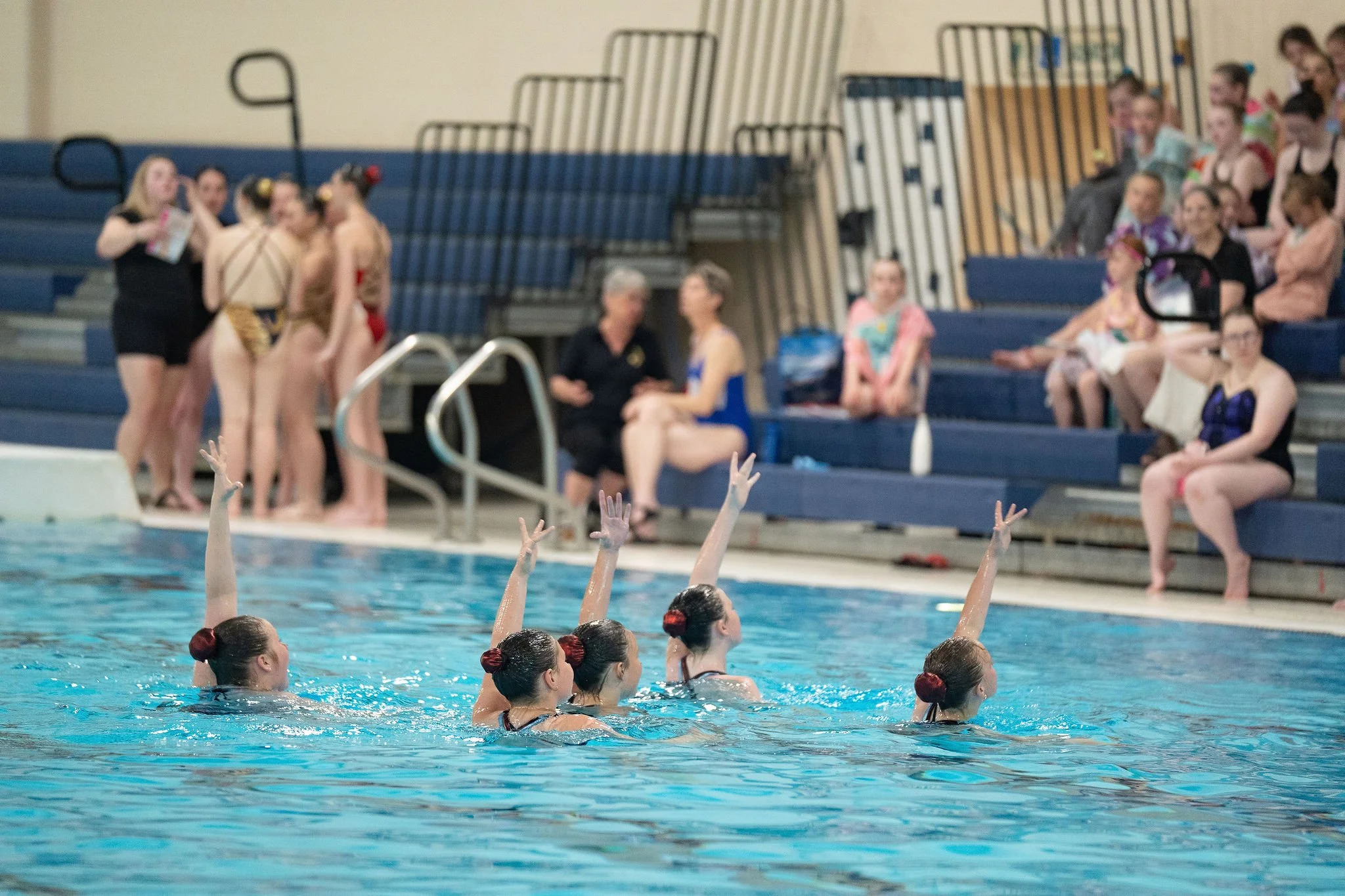 Swimmers with red hair accessories in the water during a swim meet, spectators seated on blue bleachers in the background.