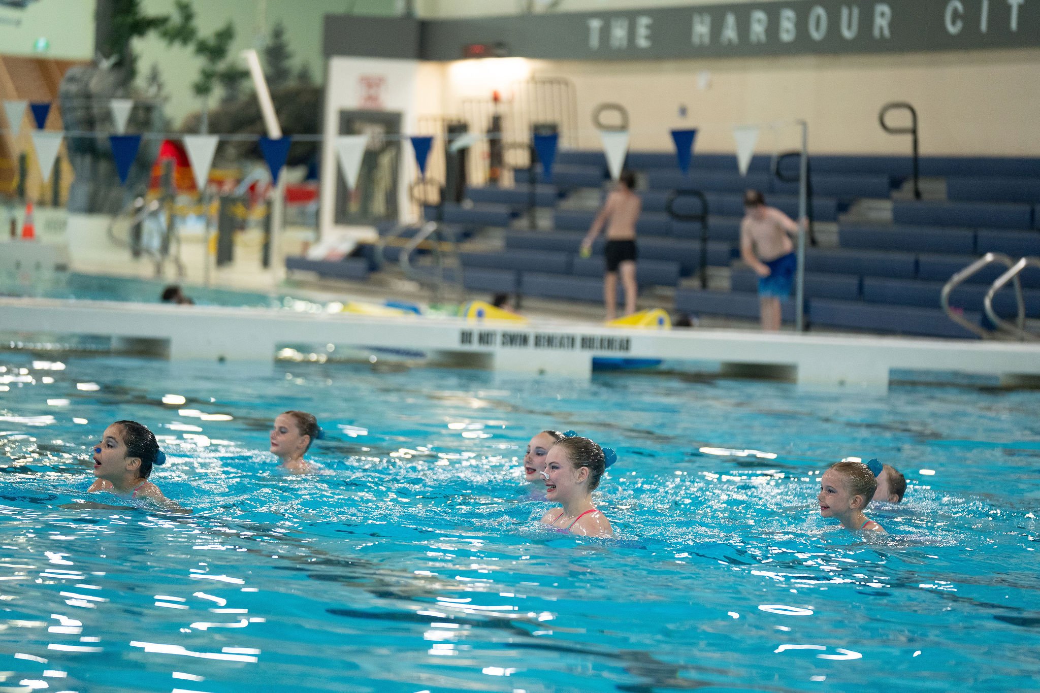 young athletes performing in the pool, with bleachers in the background.
