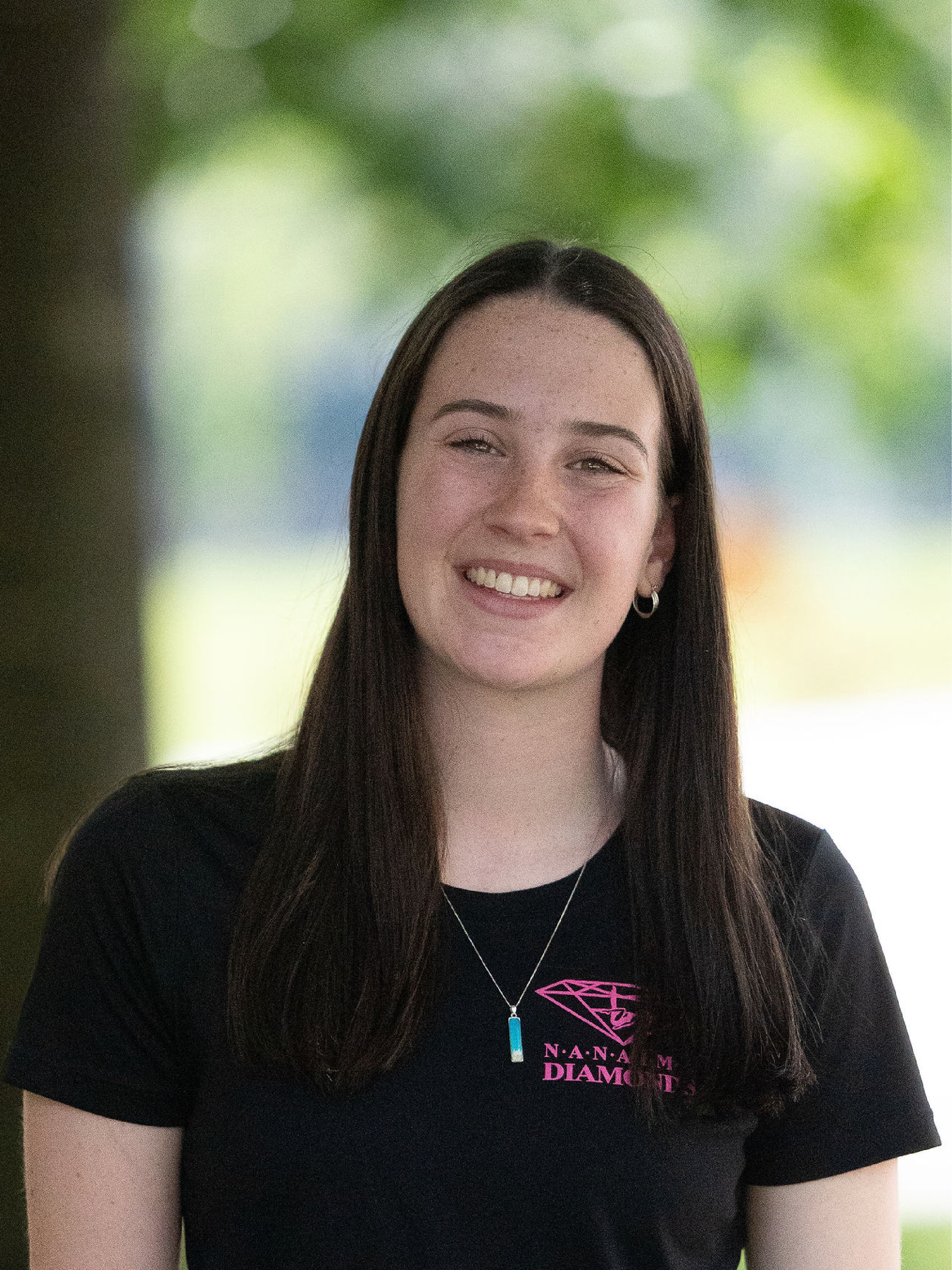 Coach Ruby, wearing a black shirt with pink nanaimo diamonds logo, standing in front of a brick wall.