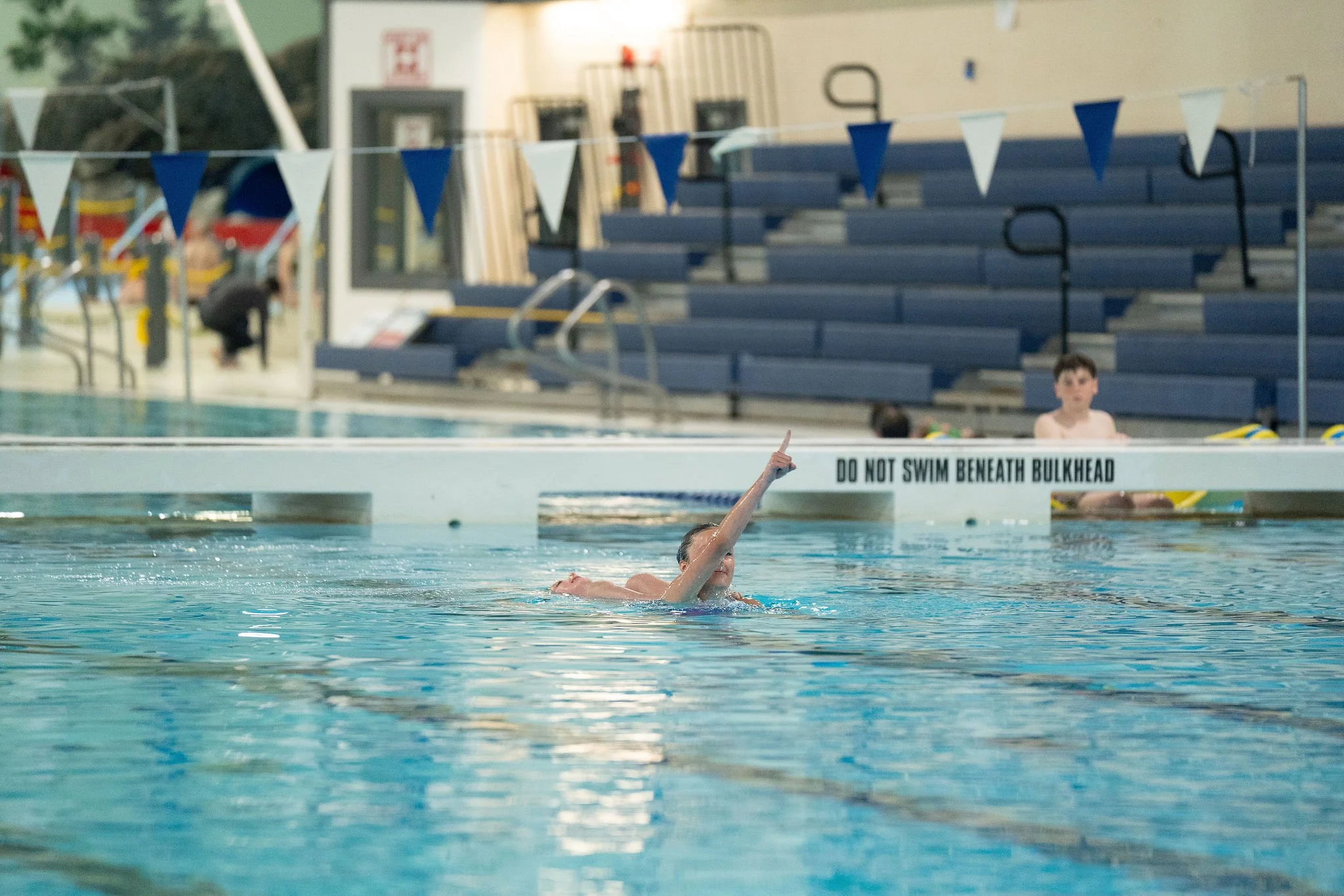 Swimmer performing in a swimming pool with blue lane dividers and bleachers in the background.