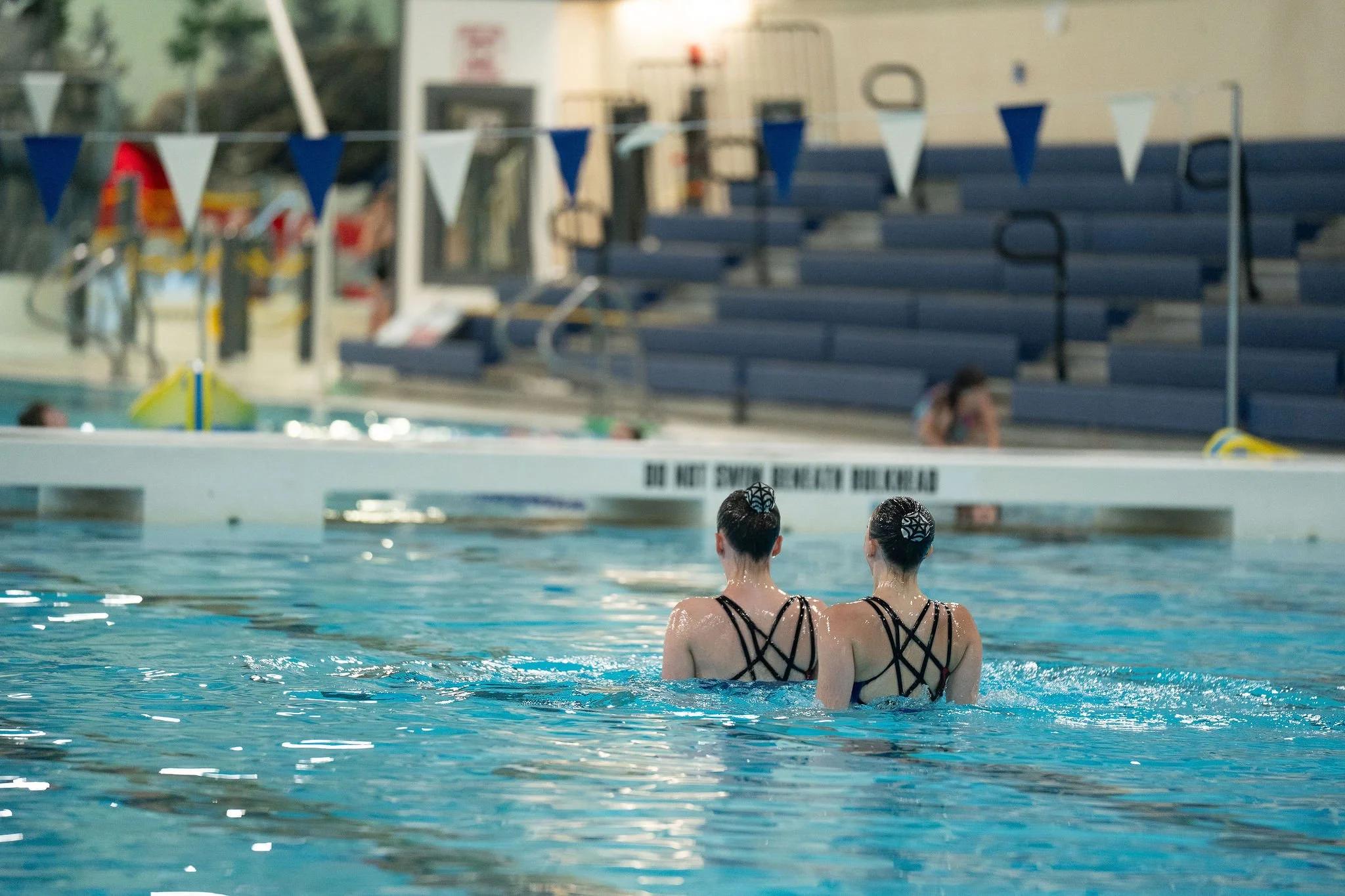 Two swimmers in competition swimsuits are performing in a swimming pool, facing away, with their hair in buns. Behind them are blue bleachers and a swimming pool with lane markings.