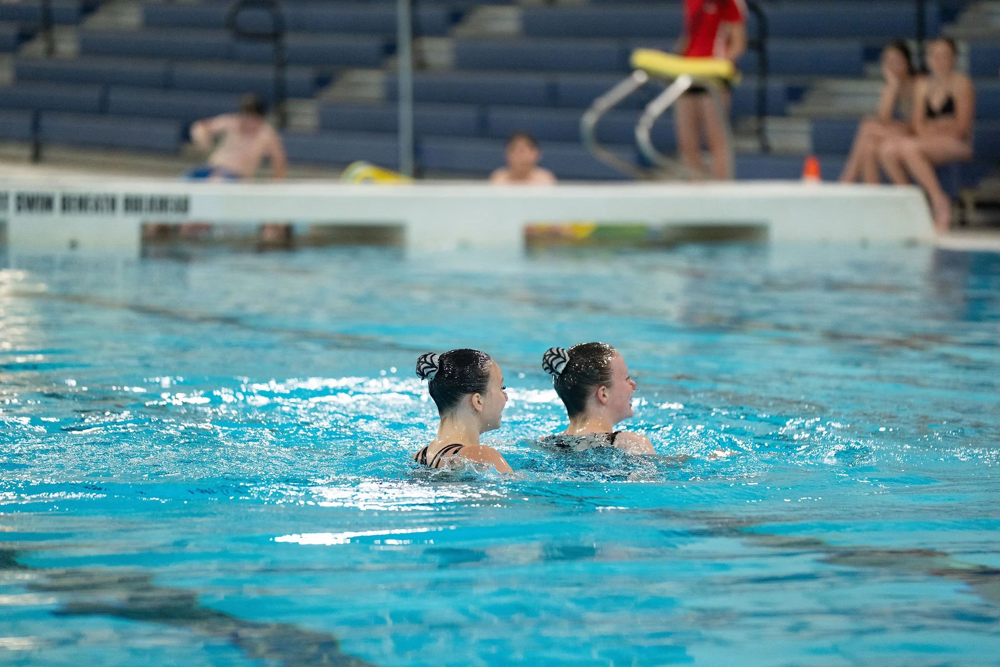 Two synchronized swimmers in a swimming pool with their hair tied up, facing forward, and wearing competition swimsuits.