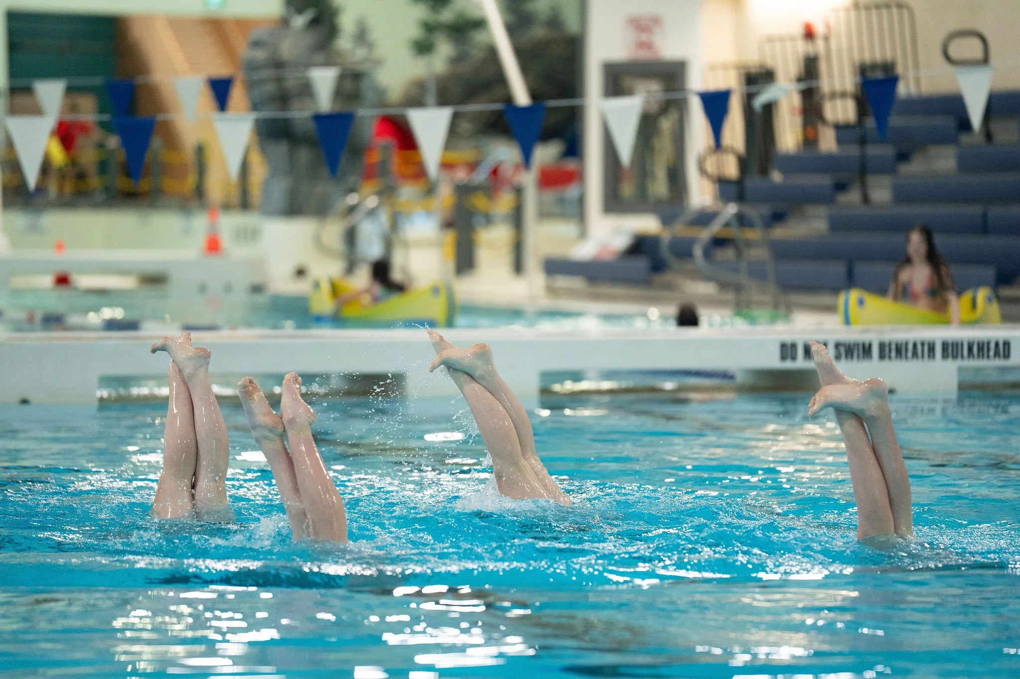 Swimming pool with multiple synchronized swimmers' legs and feet raised above the water, with a blurred background of poolside area and seating.