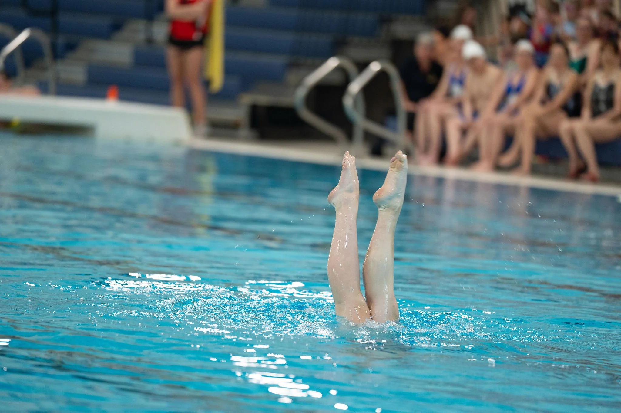 Swimmer is upside down in the pool, with legs and feet exposed out of the water, and other swimmers sitting on the pool deck watching in the background.