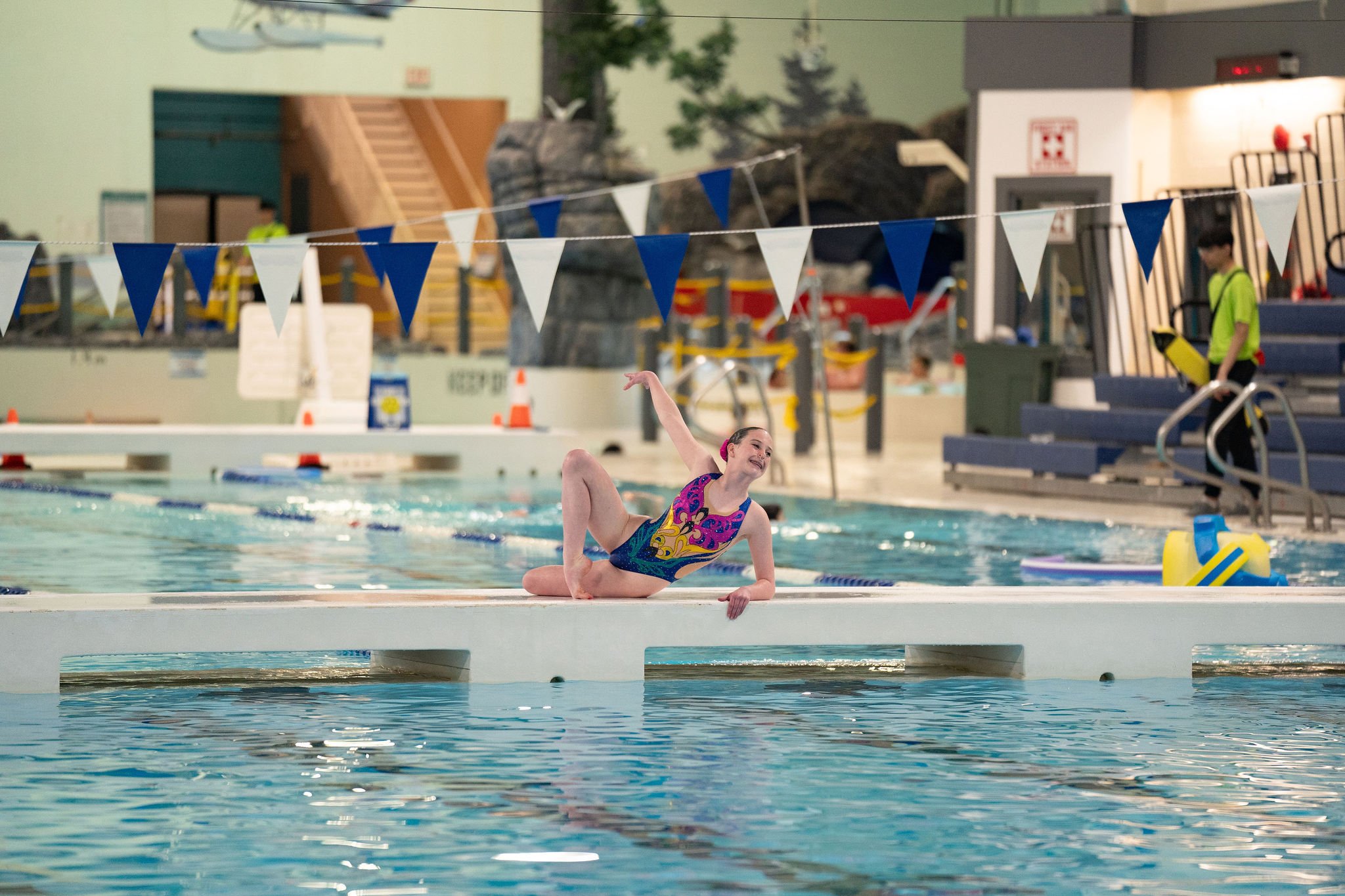 A young girl in a colorful swimsuit performing a gymnastics move on a poolside, with a swimming pool and pool equipment in the background.
