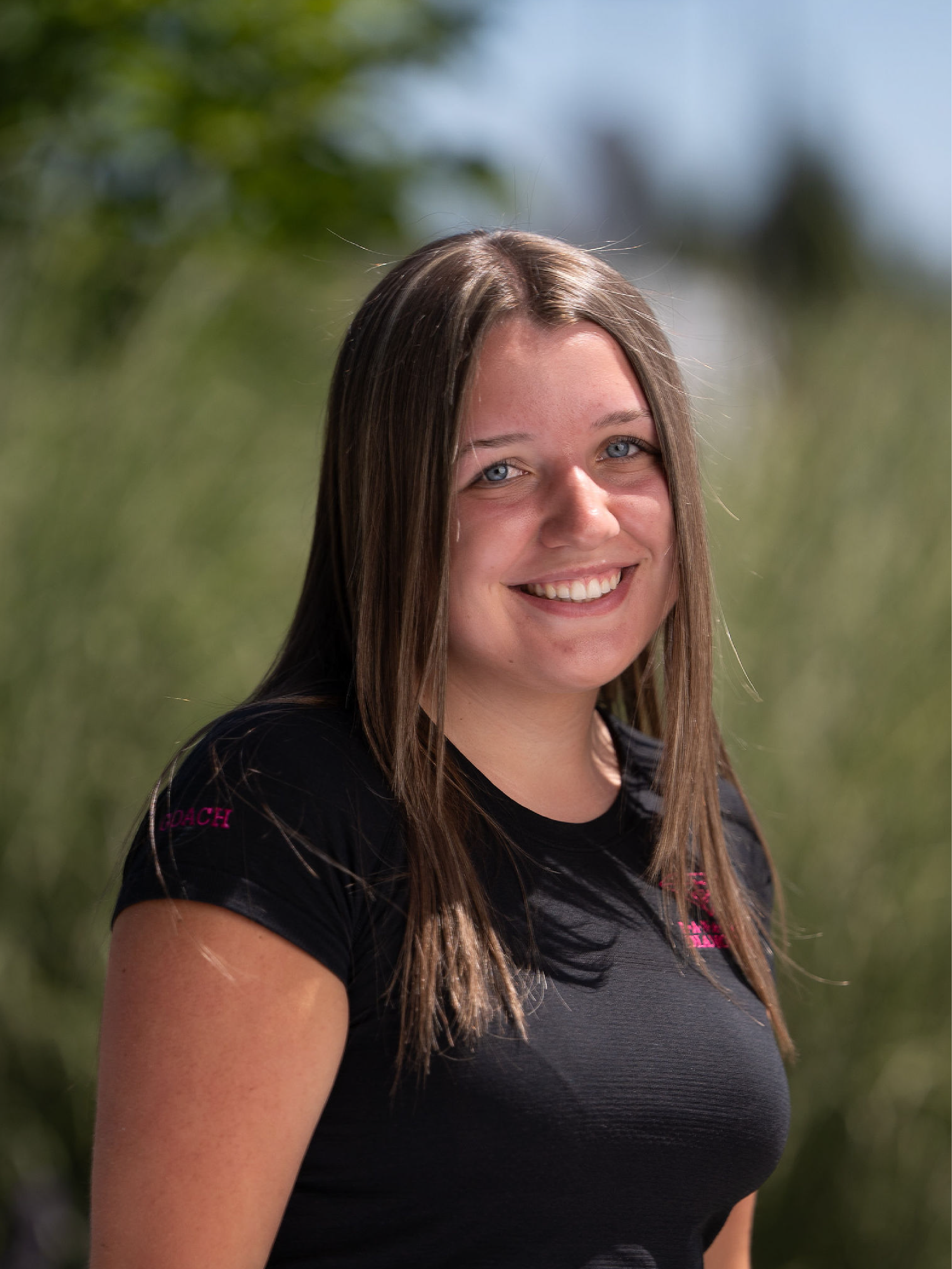 Coach Ryleigh, wearing a black shirt with pink nanaimo diamonds logo, standing in front of a brick wall.