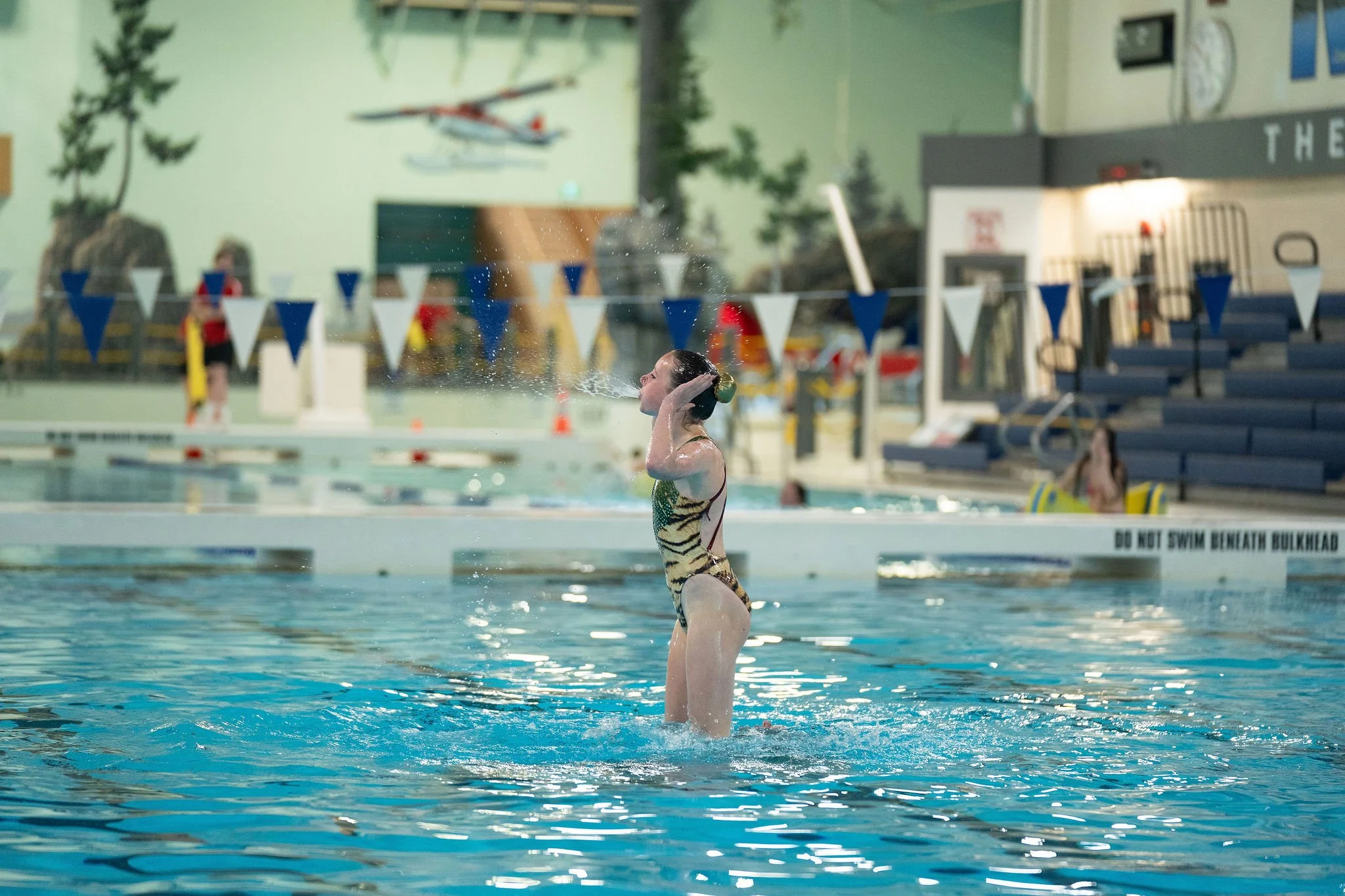 A female synchronized swimmer standing in a swimming pool with her arms raised and adjusting her swim cap, with a diving board and other swimmers in the background.