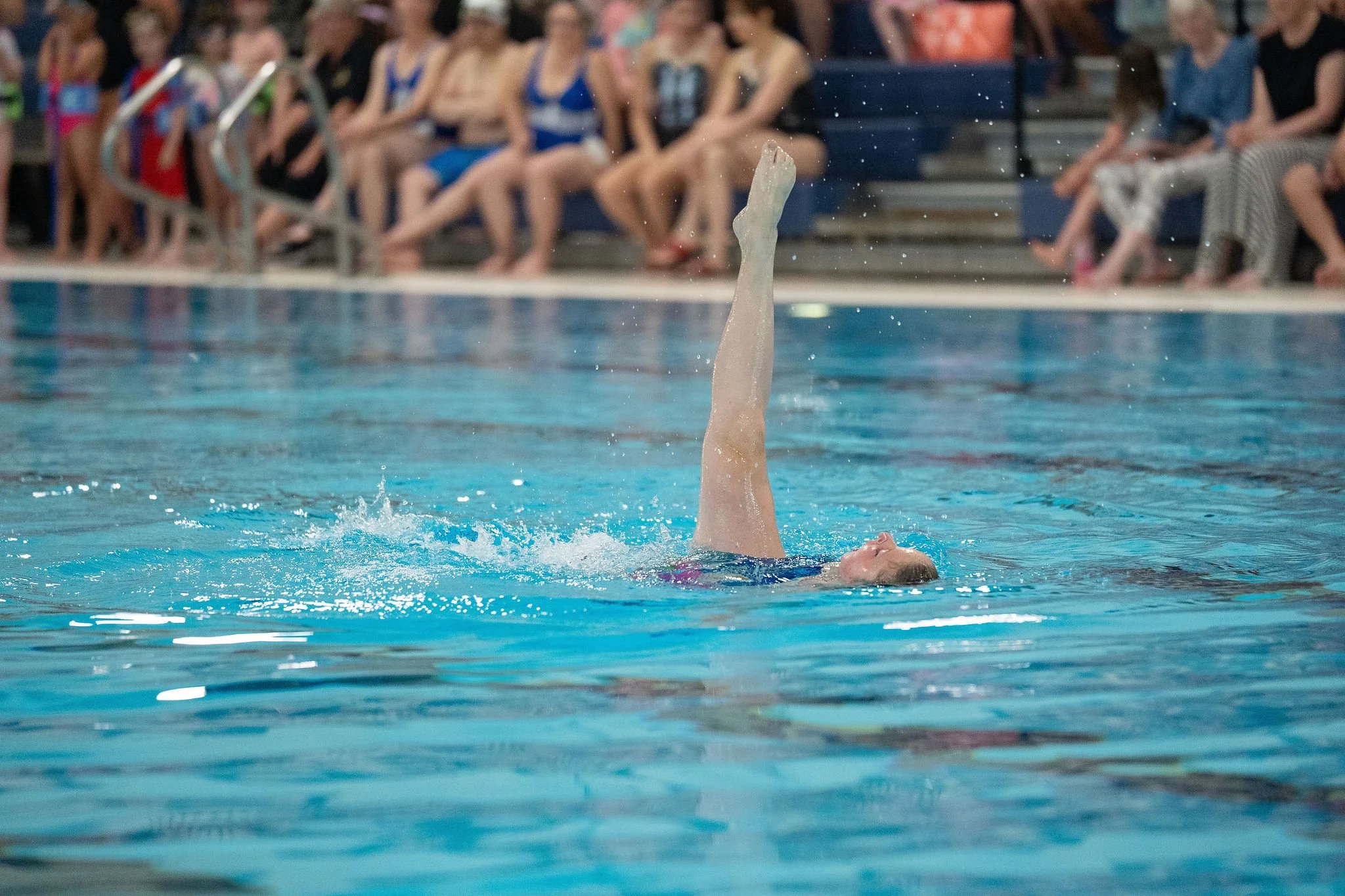A synchronized swimmer performs a ballet leg in the pool, while spectators watch from the bleachers in the background.