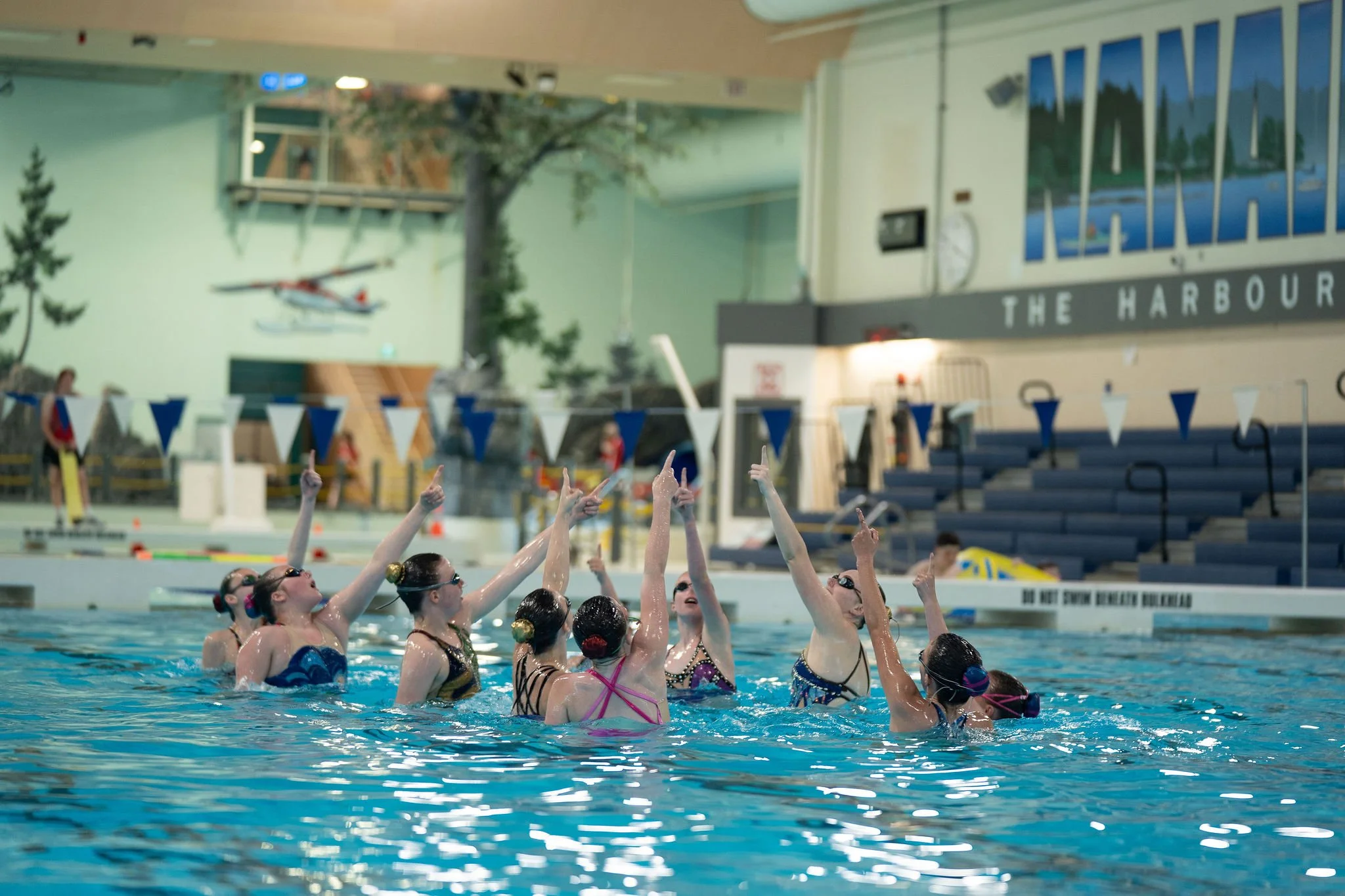 Swimmers in a swimming pool raising their hands, with an indoor aquatic center in the background, including a mural, seating area, and a visible sign with safety instructions.