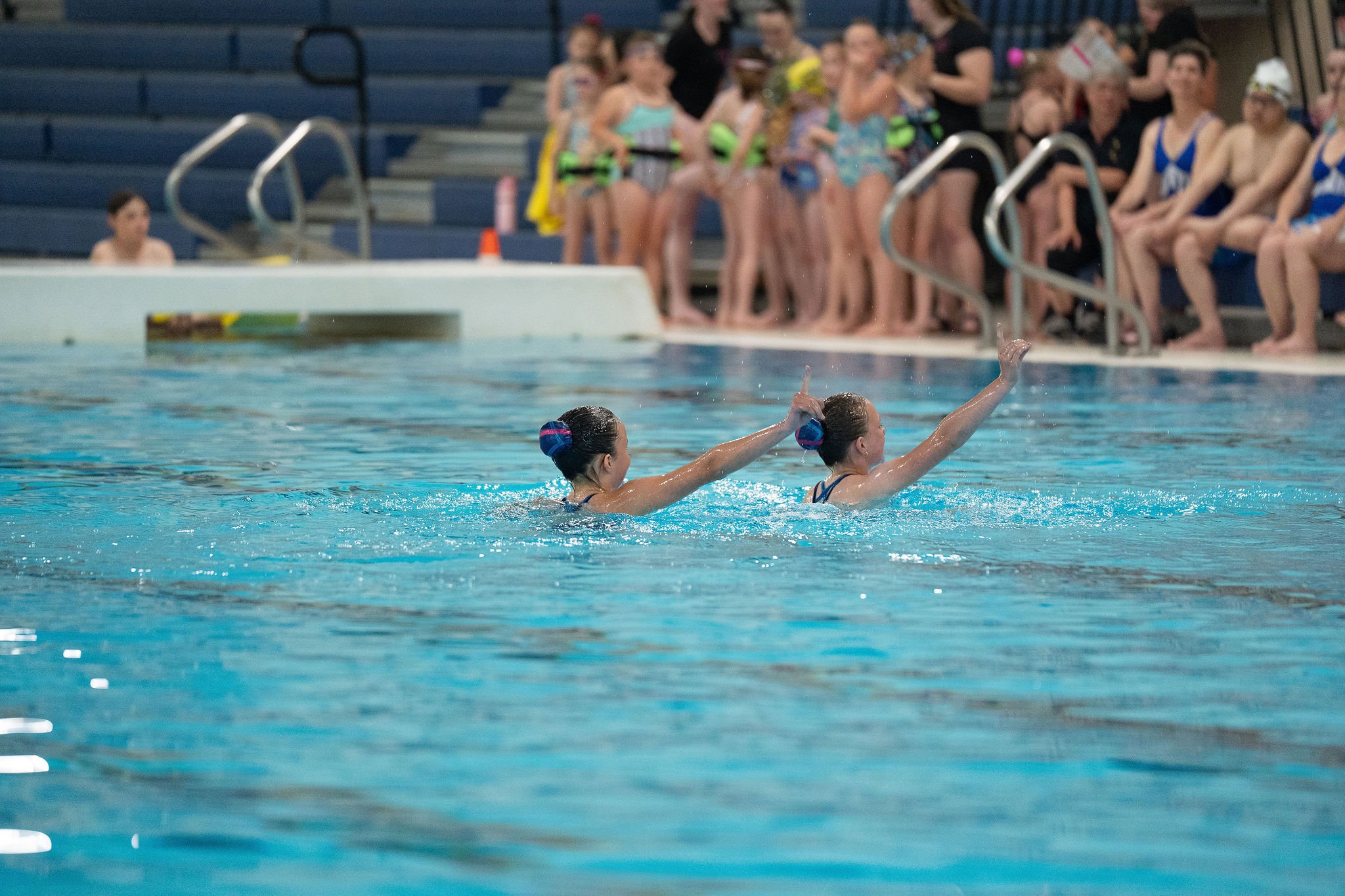 Swimmers performing synchronized swimming routine in a pool with spectators watching from the stands.