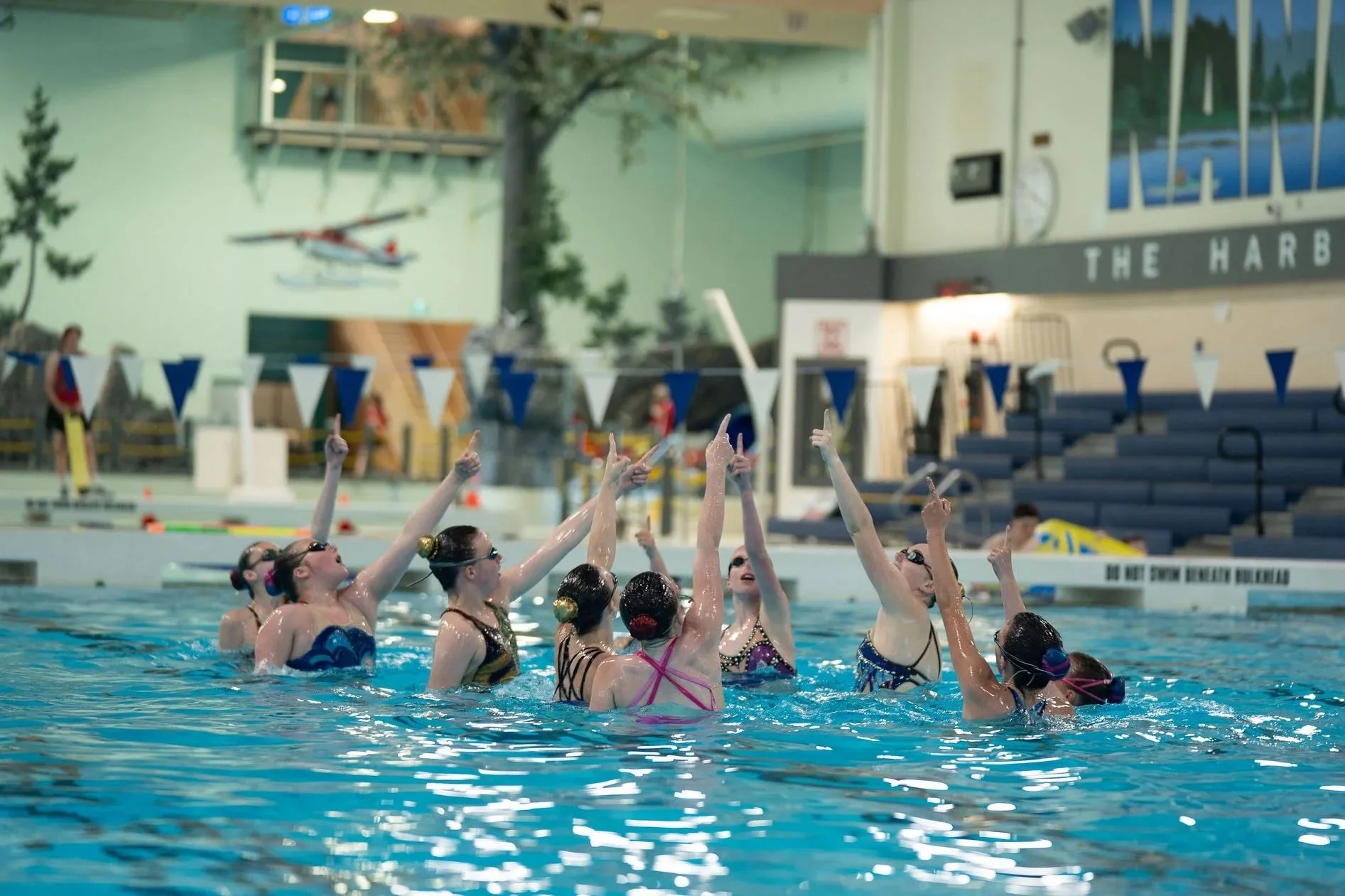 Swimmers peforming in a swimming pool raising their hands, during a swimming showcase, with a large indoor aquatic facility in the background.