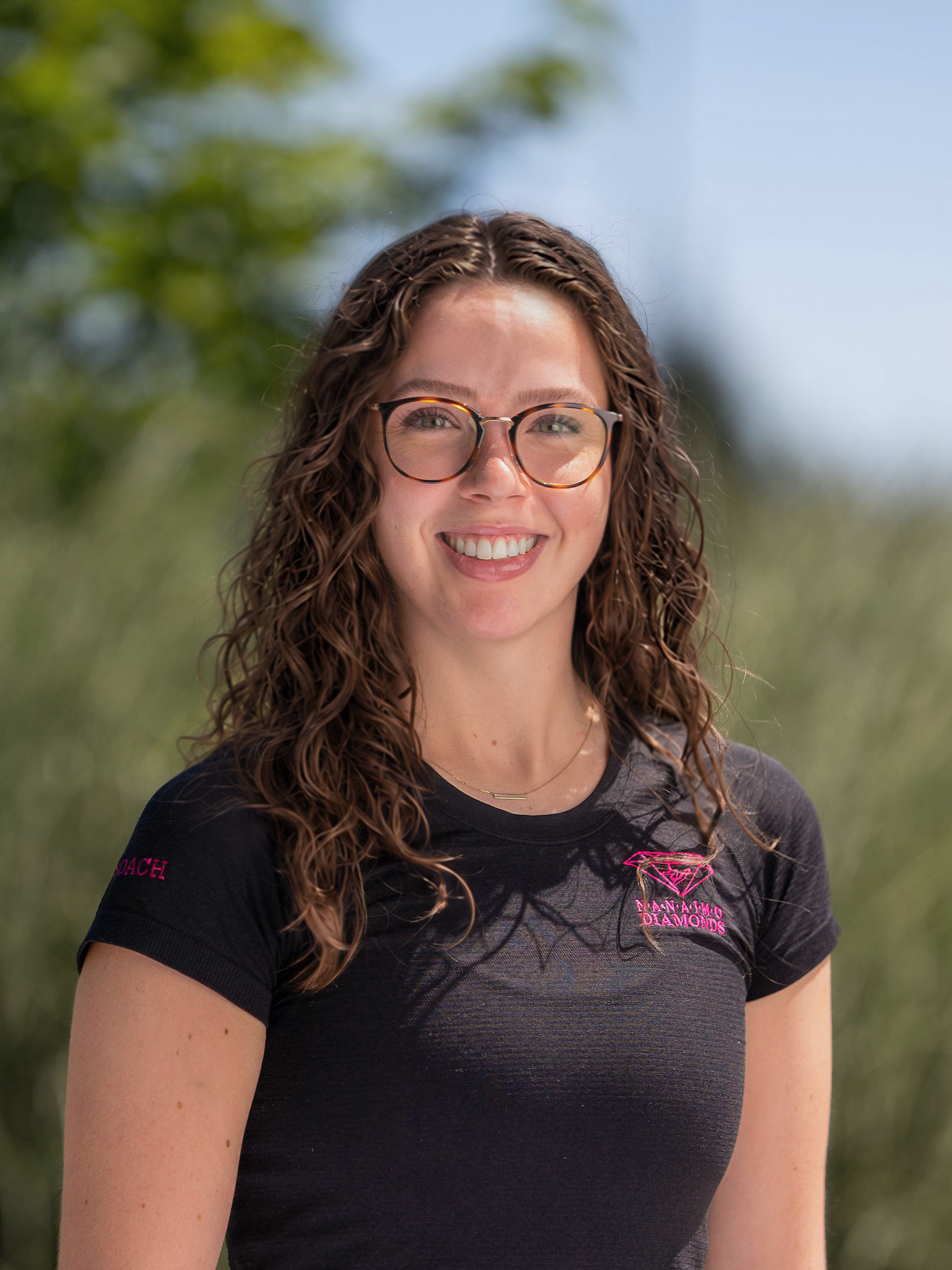 Coach Jess, wearing glasses, a black shirt with pink nanaimo diamonds logo, standing in front of a brick wall.