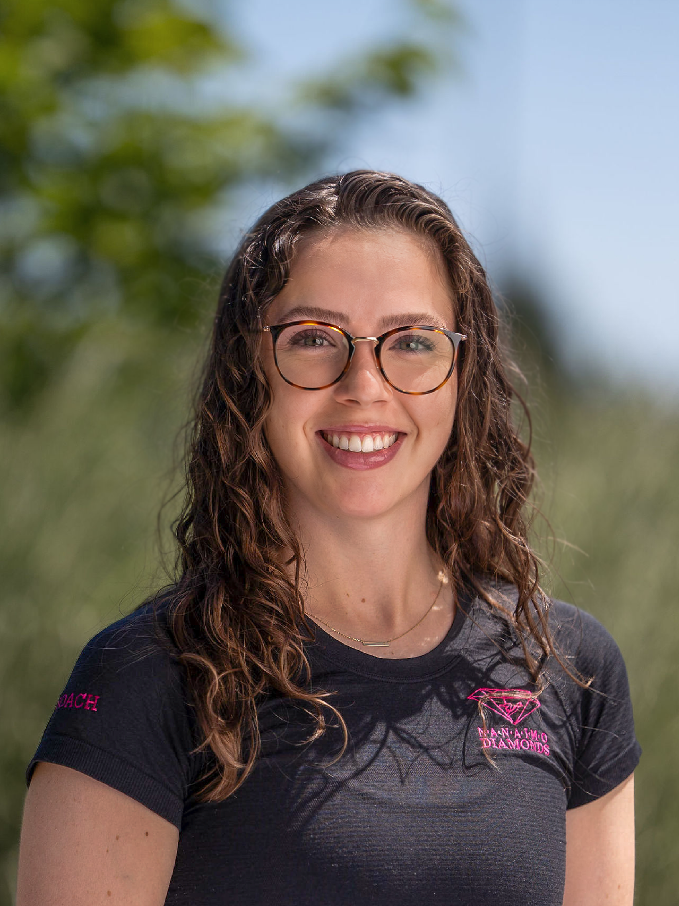 Coach Jess, wearing glasses, a black shirt with pink nanaimo diamonds logo, standing in front of a brick wall.