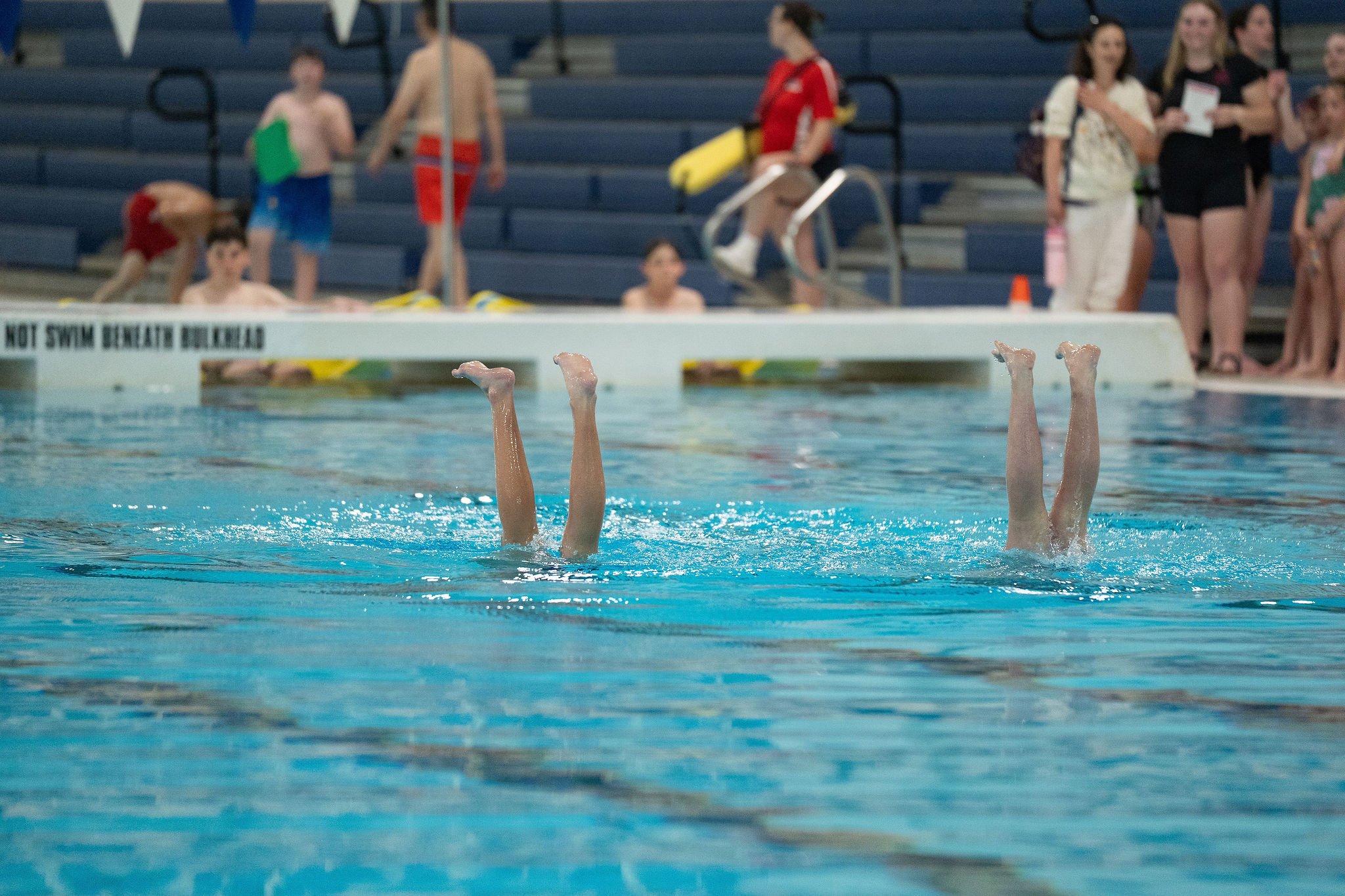 Swimmers' legs in the air at a swimming pool during a competition, with people observing in the background.