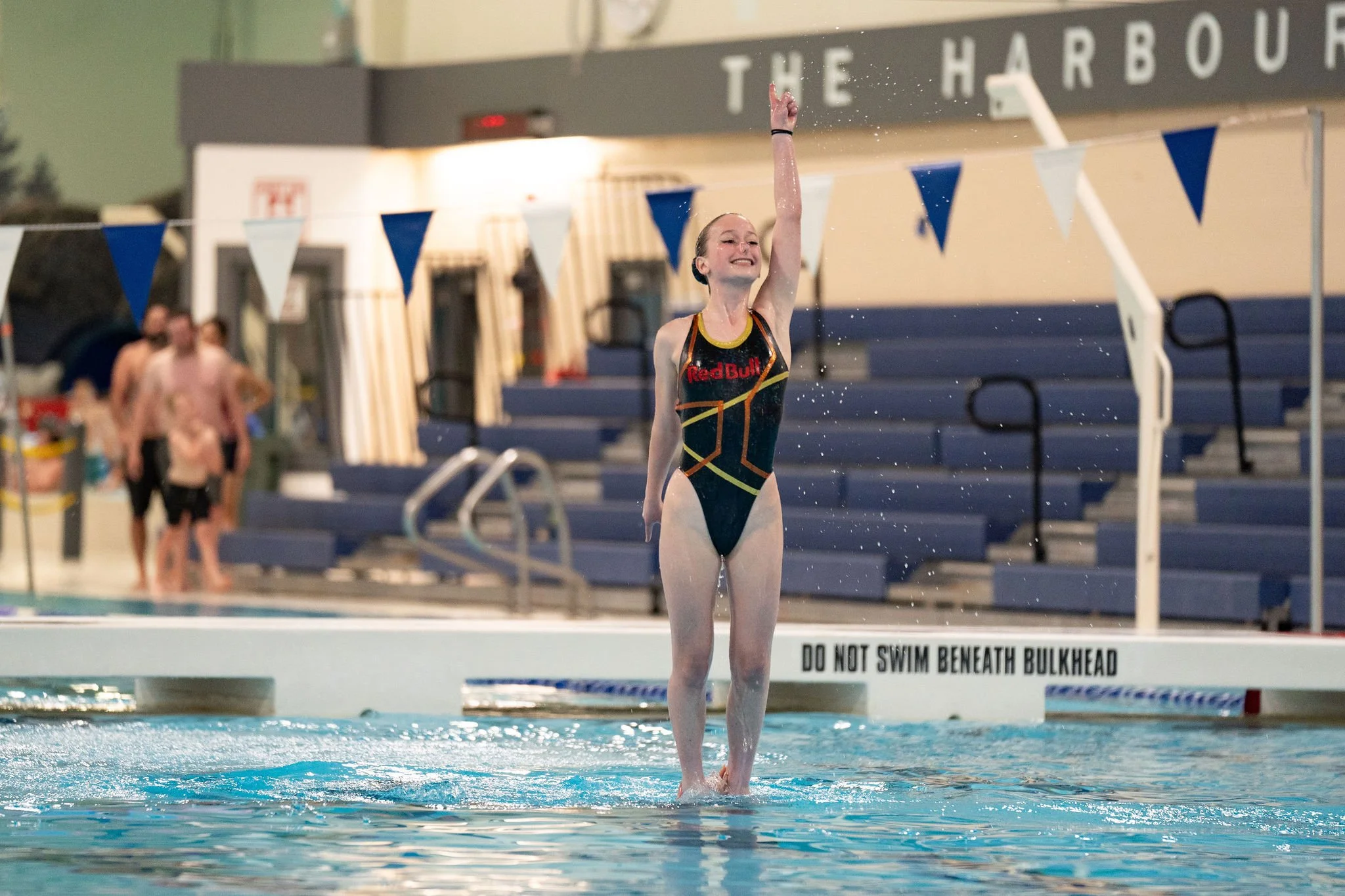 A young woman in a black swimsuit with yellow and red accents, smiling and raising her right arm in the air, standing on an acrobatic during a competition.
