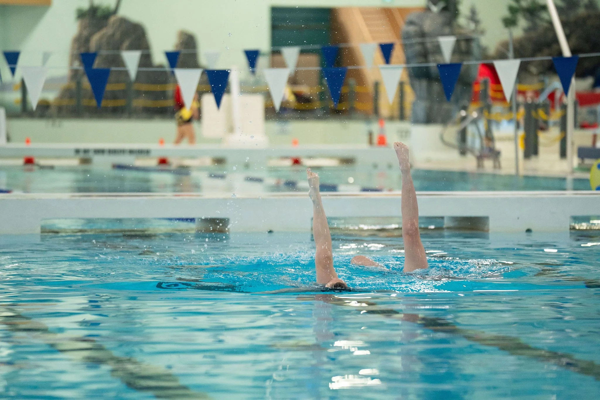 Two synchronized swimmers perform ballet legs in the pool, while spectators watch from the bleachers in the background.