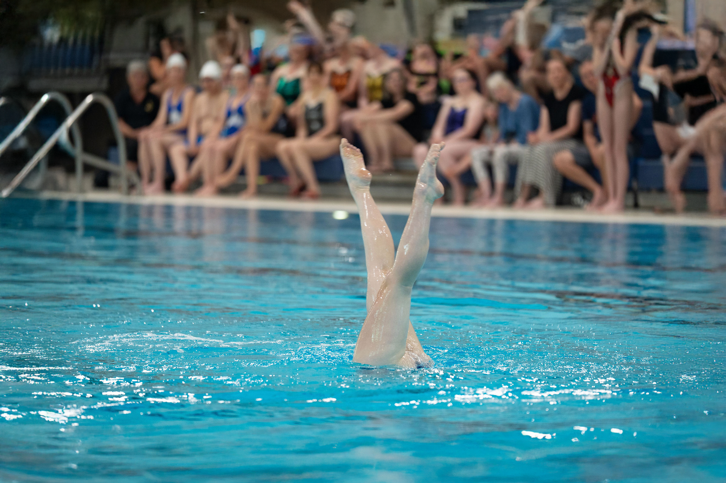 Swimmer is upside down in the pool, with legs and feet exposed out of the water, and other swimmers sitting on the pool deck watching in the background.