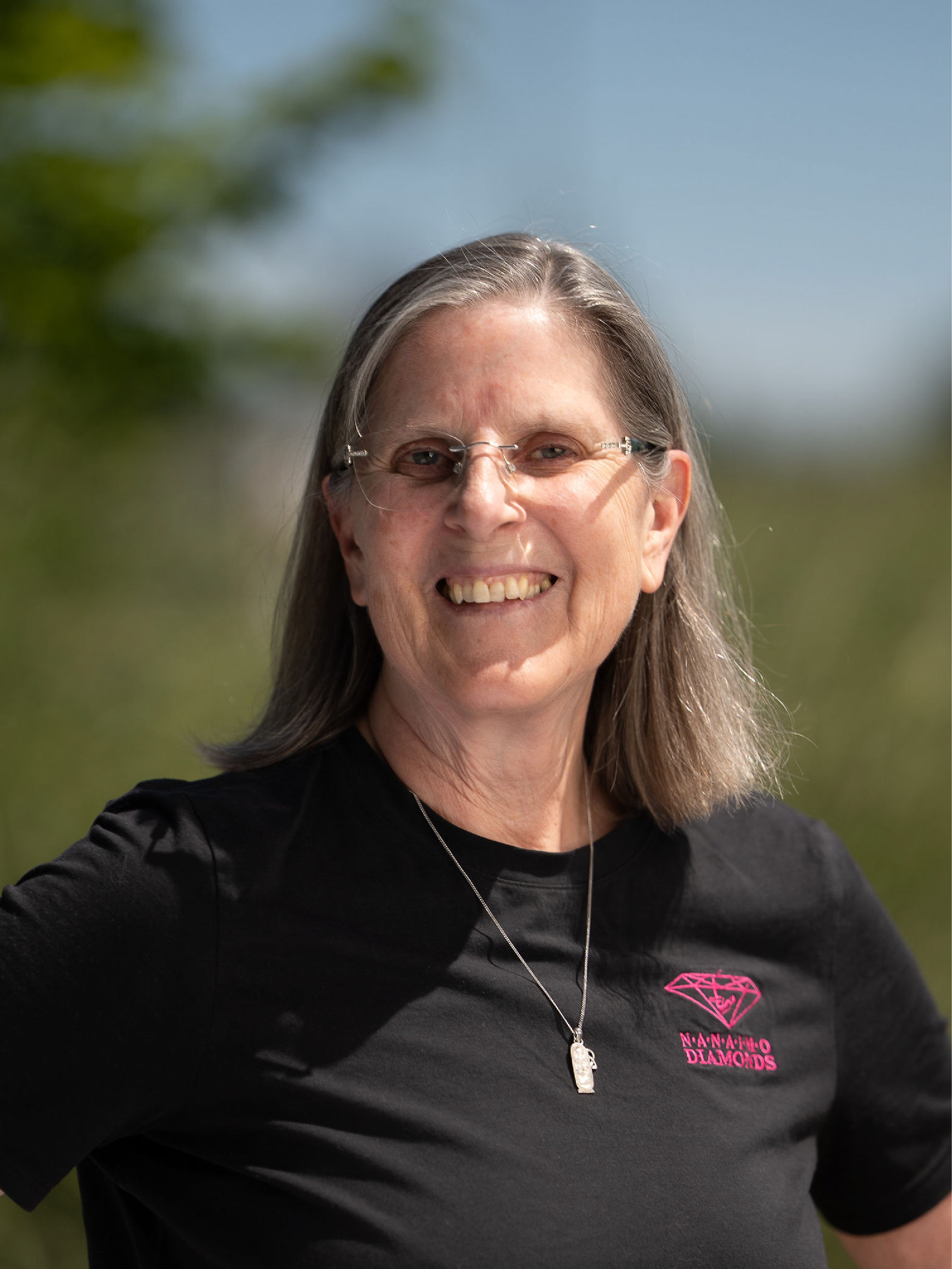 Catharine Edwards our Head Coach, wearing red glasses and a black shirt with pink nanaimo diamonds logo, standing in front of a brick wall.