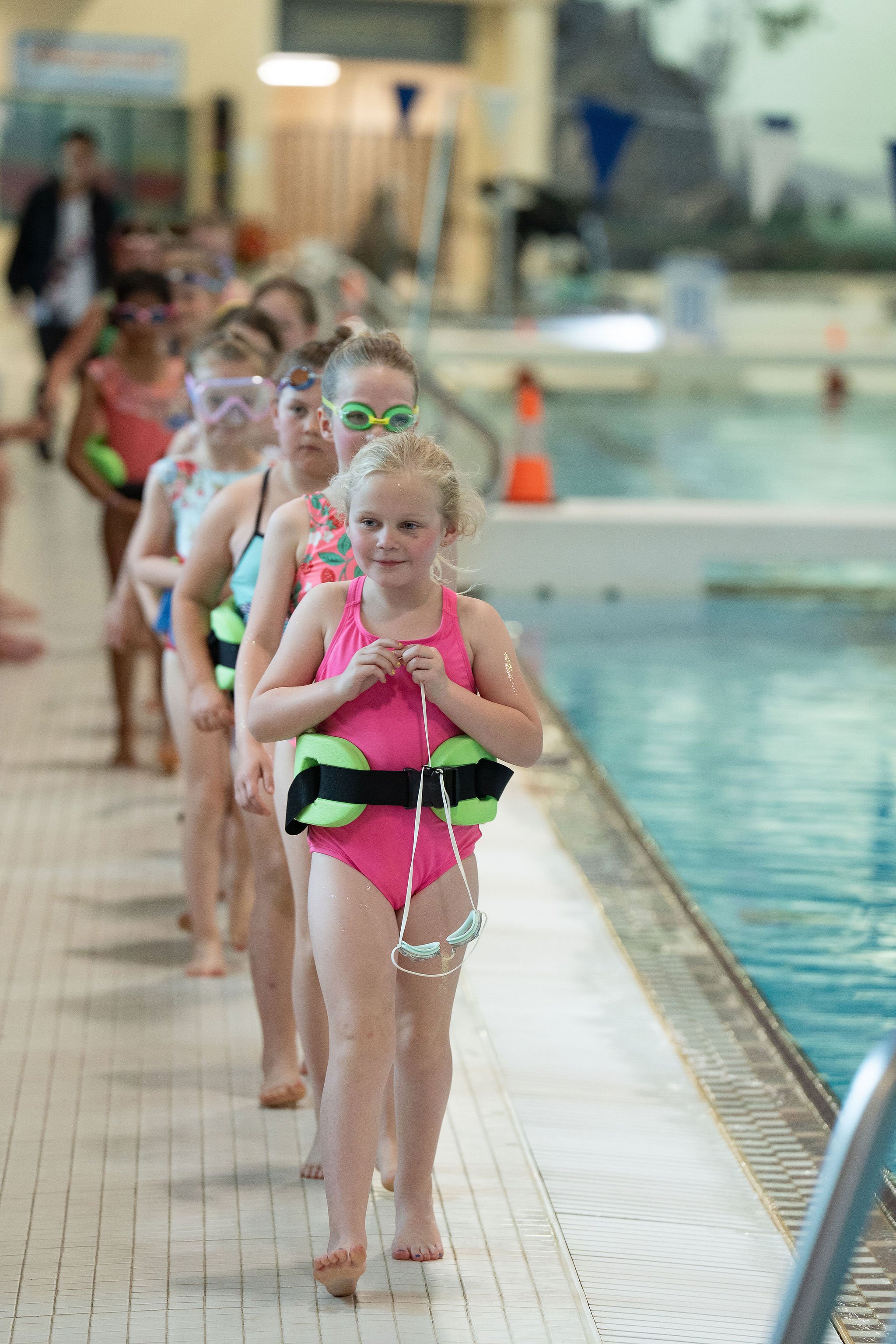 Children in swimsuits and goggles standing in a line by an indoor swimming pool.