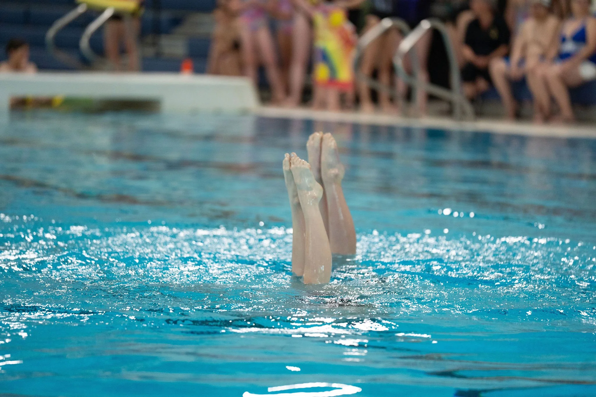 Two athletes with their legs and feet visible as they perform in the water. In the background, there are several people sitting on the poolside chairs and standing, some wearing swimsuits and swimming caps.