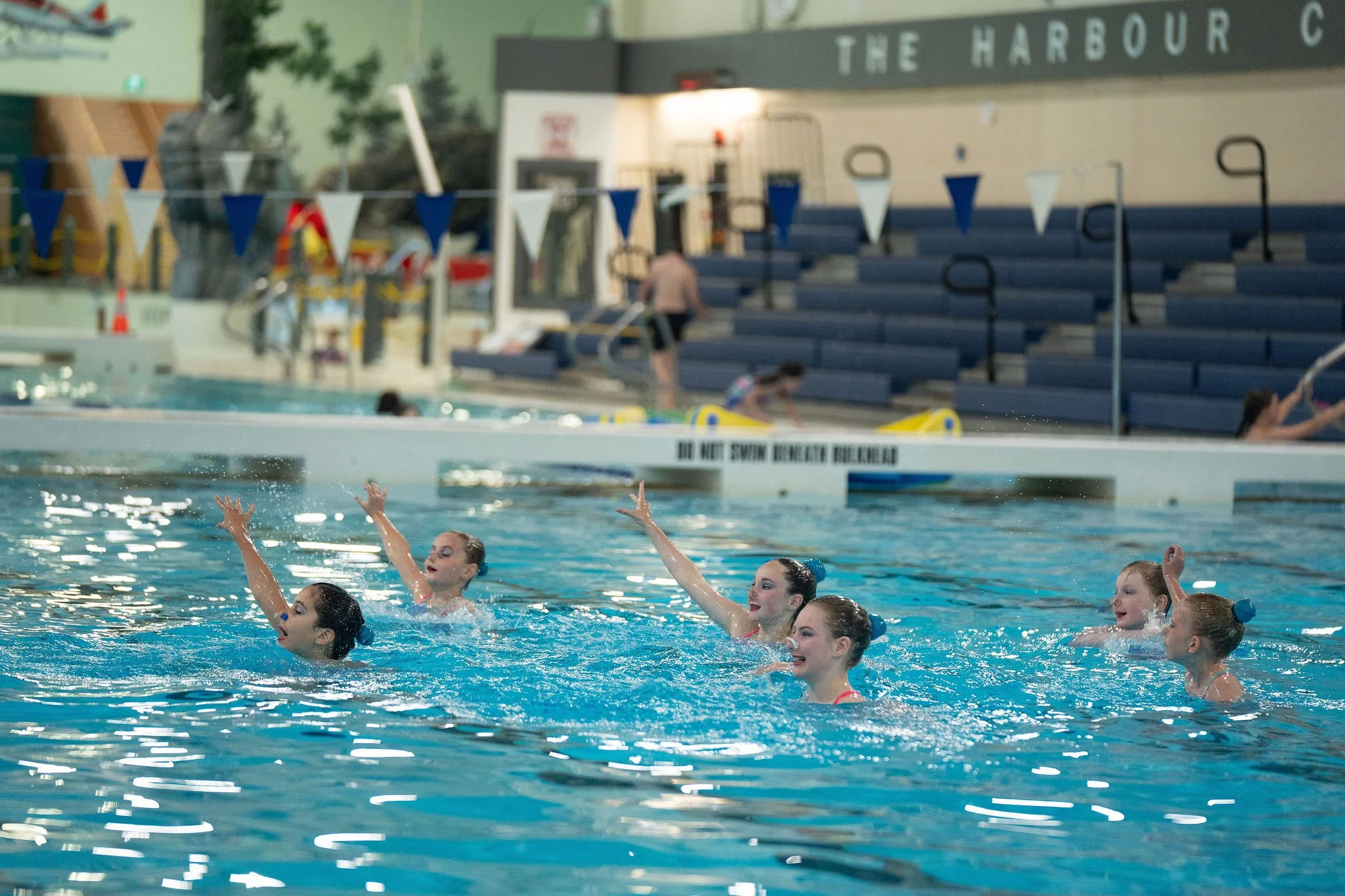 Young athletes performing in the pool, with bleachers in the background.